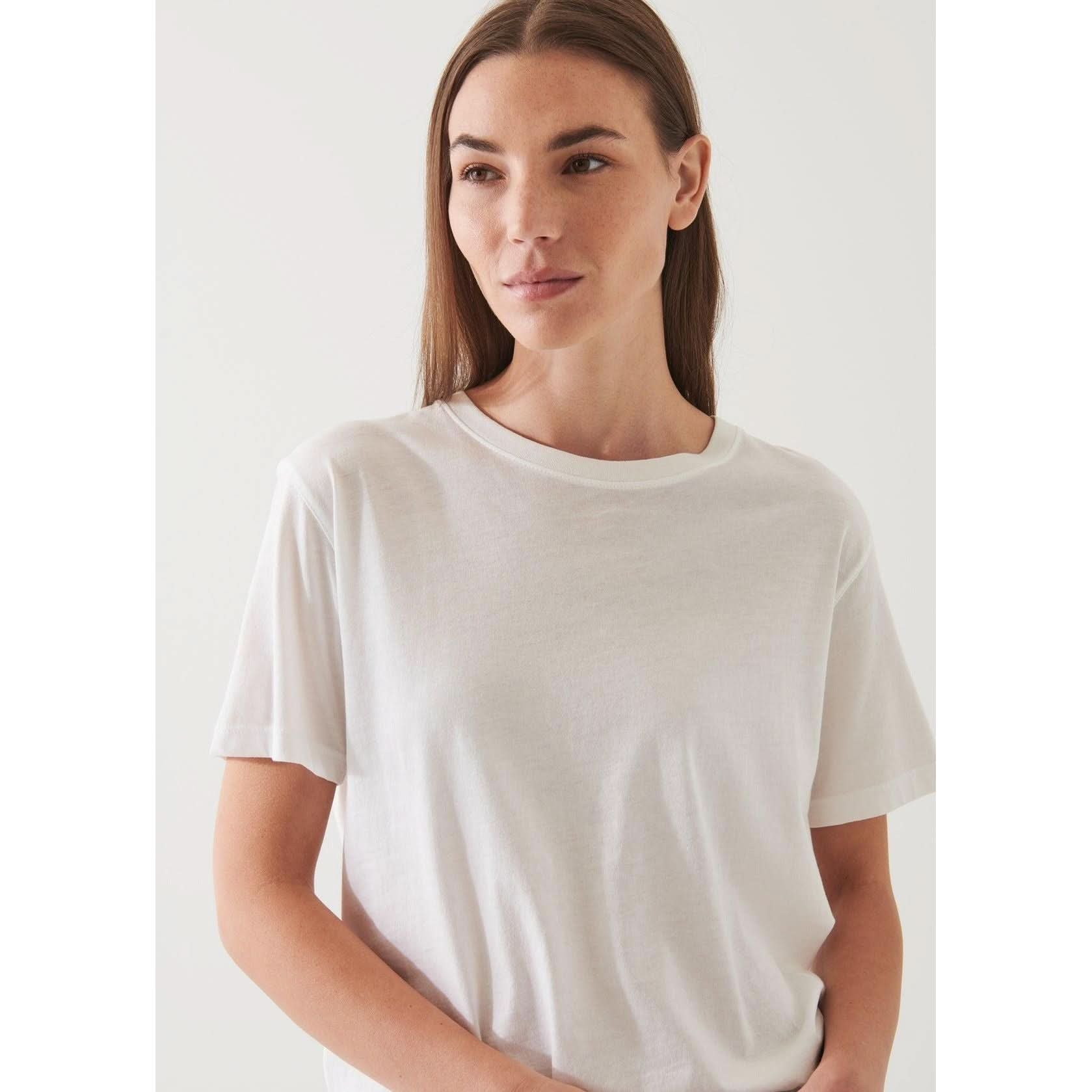 A woman with long brown hair stands against a plain light-colored background, wearing an oversized fit, Patrick Assaraf SS Iconic Boyfriend Crew White made of soft pima cotton. She has a calm expression and is posing with one hand resting at her side.