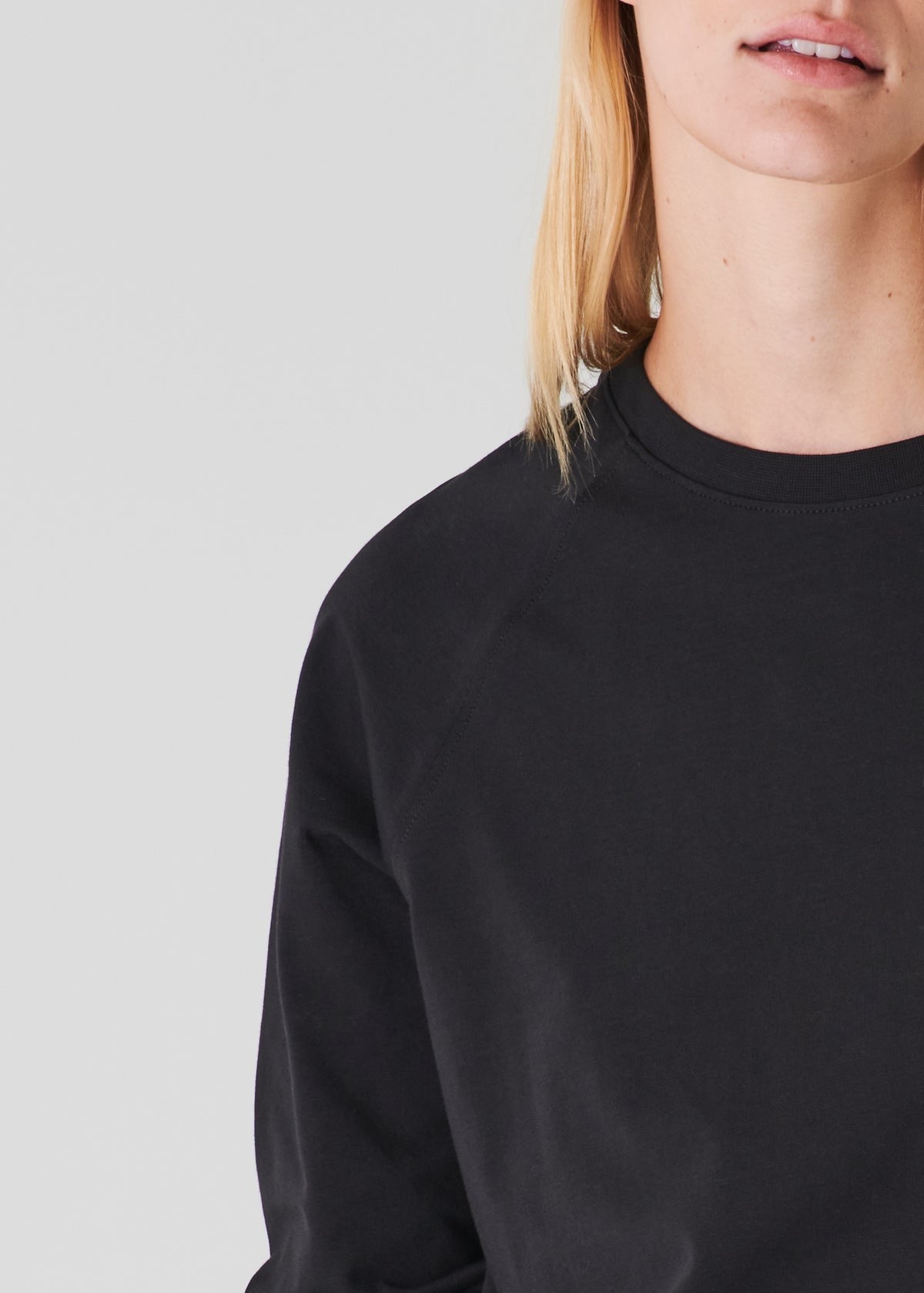 Close-up of a person with long, blonde hair, visible from the neck to the shoulder, wearing a relaxed fit Patrick Assaraf Pima Cotton Stretched Oversized Sweatshirt in black. The background is a pale, solid color. Only the lower part of their face is in frame, not showing their eyes.