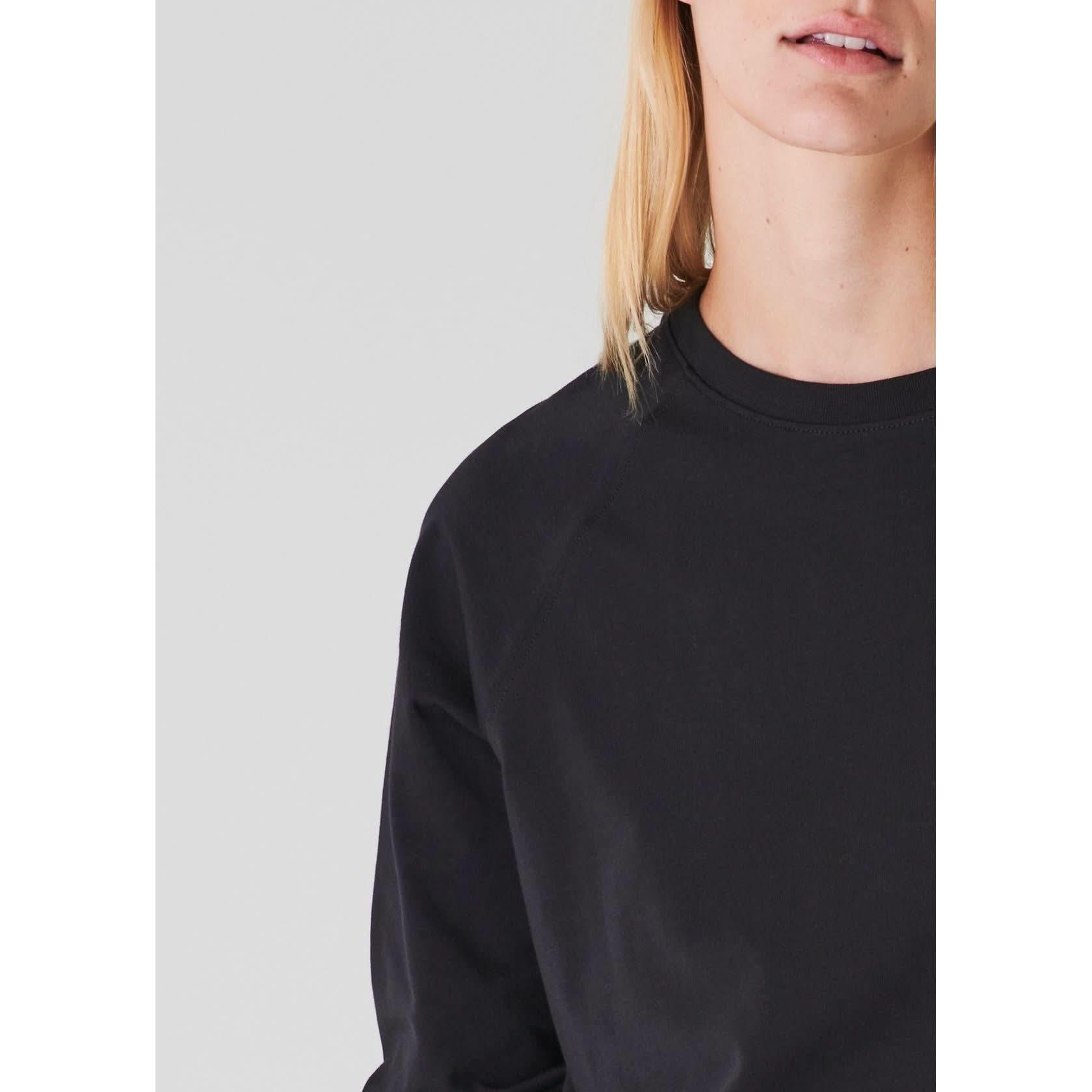 Close-up of a person with long, blonde hair, visible from the neck to the shoulder, wearing a relaxed fit Patrick Assaraf Pima Cotton Stretched Oversized Sweatshirt in black. The background is a pale, solid color. Only the lower part of their face is in frame, not showing their eyes.