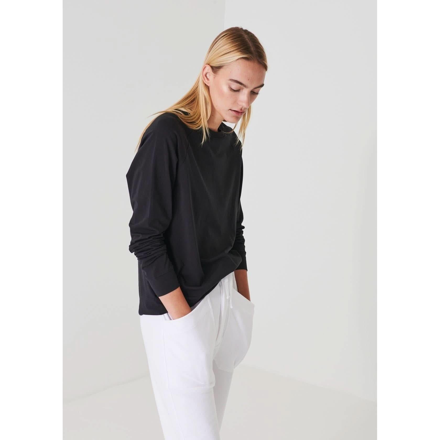 A person with long, straight blonde hair is wearing a Patrick Assaraf Pima Cotton Stretched Oversized Sweatshirt in black and white pants. They have their hands in their pockets and are looking downward thoughtfully in a minimalistic, light-colored room.