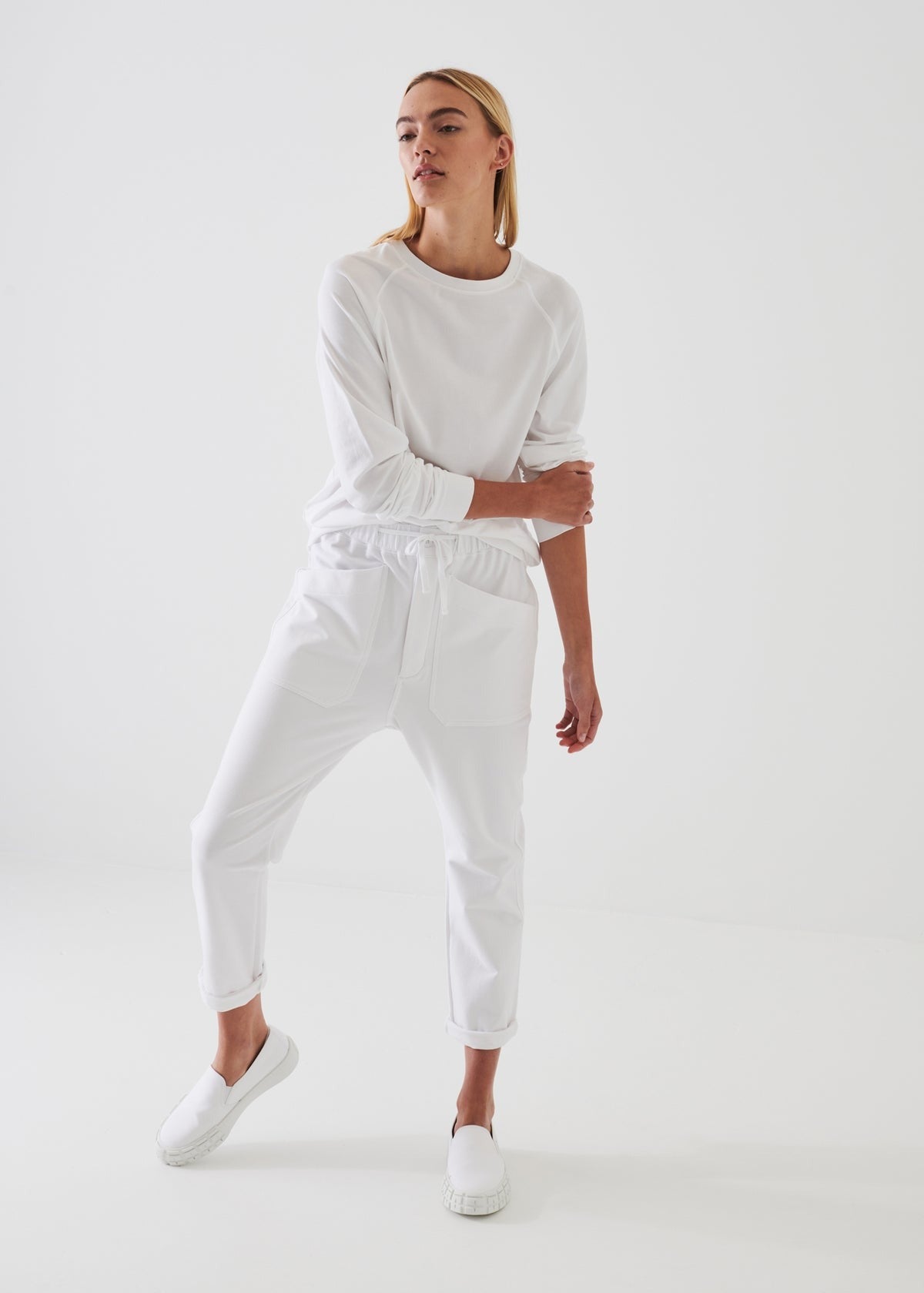 A person stands against a plain white background, dressed in a relaxed fit Patrick Assaraf Pima Cotton Stretch Oversized Sweatshirt in Chalk and drawstring pants. The individual is wearing white slip-on shoes, with one hand resting on their elbow and looking slightly to the side.