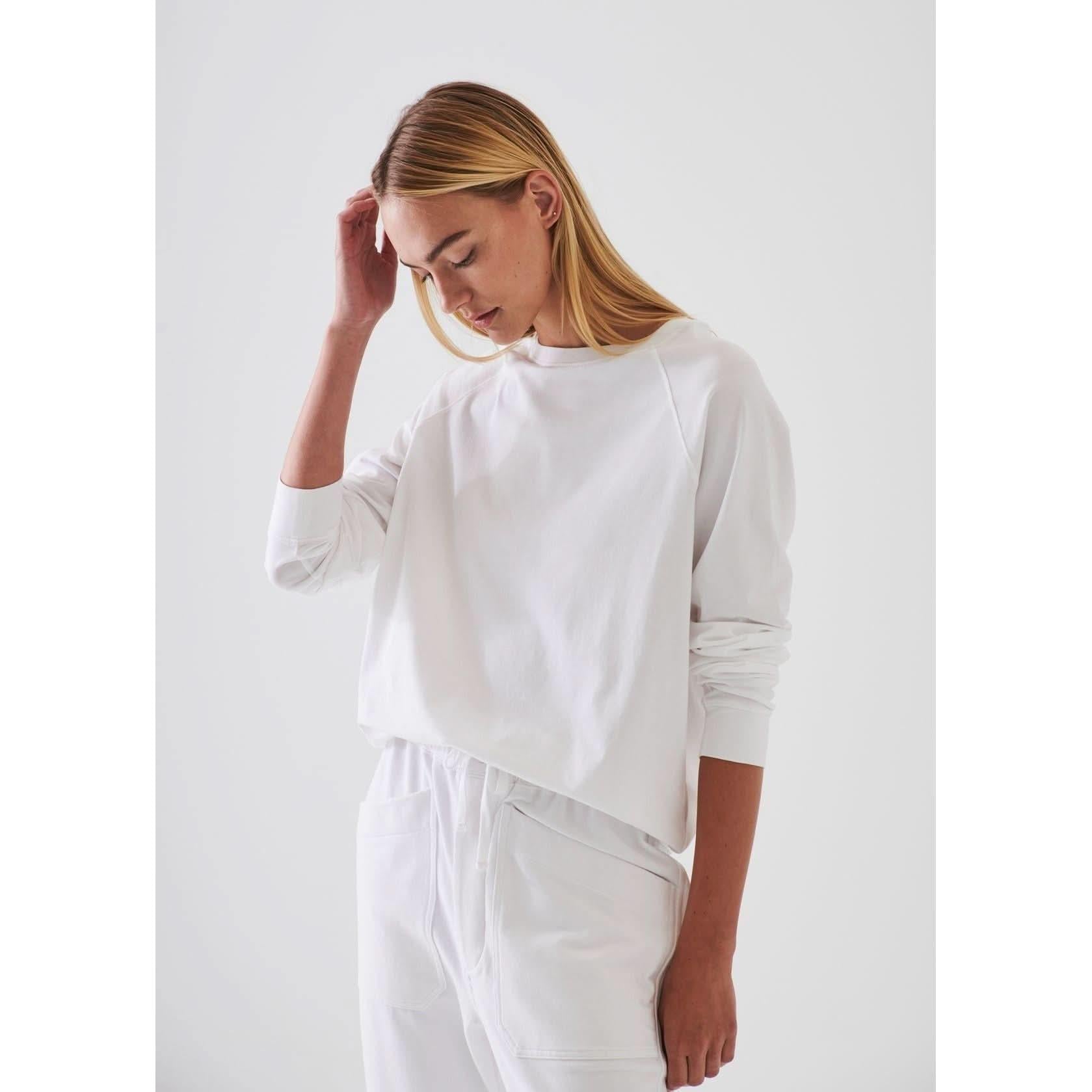 A person with long blonde hair is wearing the Patrick Assaraf Pima Cotton Stretch Oversized Sweatshirt in Chalk and white pants. They are looking down and gently touching their head with their right hand, standing against a plain white background, showcasing the relaxed fit of this Peruvian pima cotton ensemble from Patrick Assaraf.