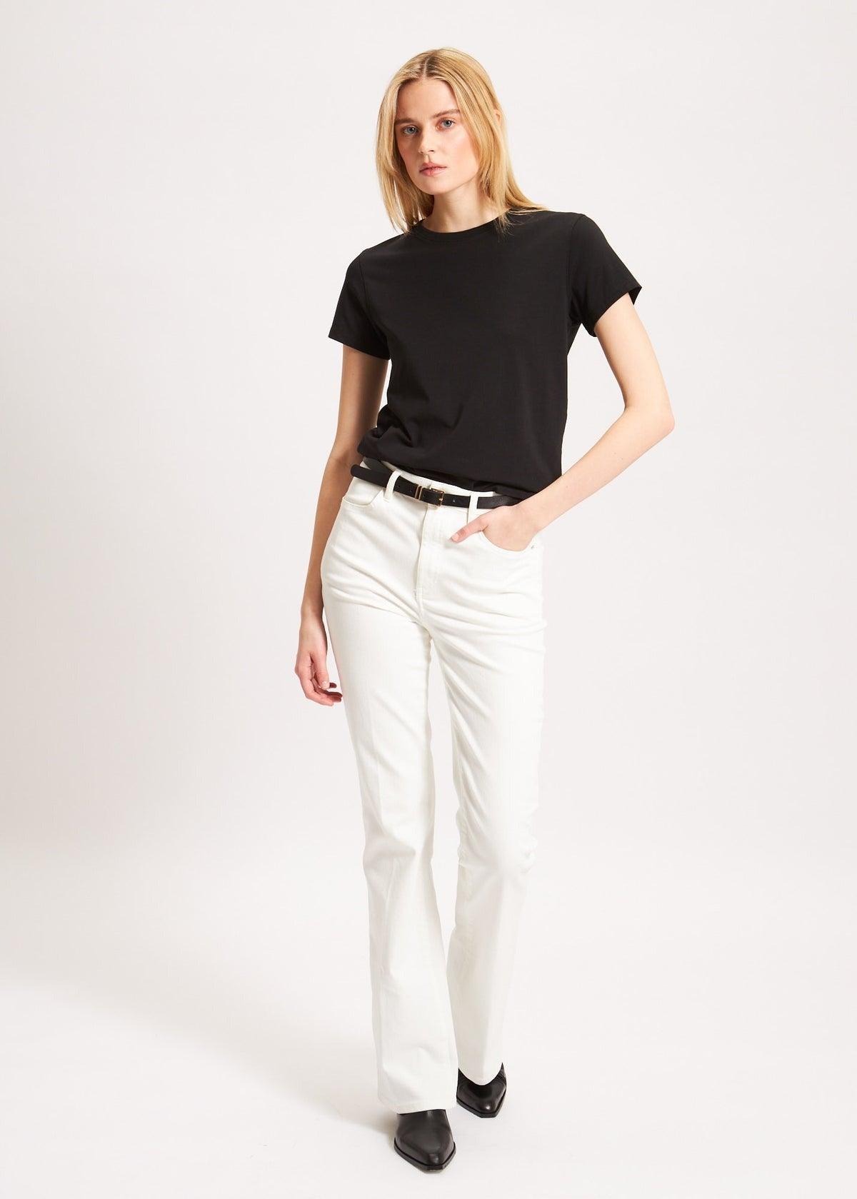 A woman with long blonde hair is standing against a plain background. She is wearing a Patrick Assaraf SS Iconic Fitted Crew Black T-shirt, crafted from luxurious Peruvian pima cotton, tucked into high-waisted, white straight-leg pants. With one hand in her pocket and black shoes on her feet, she exudes effortless style.