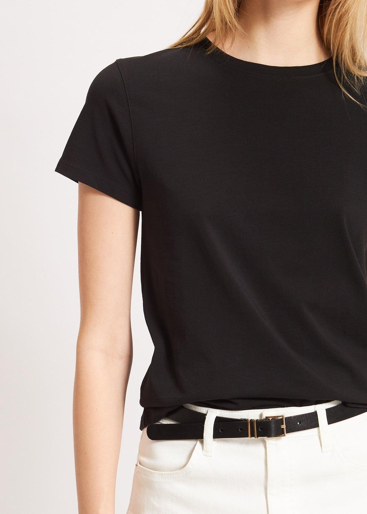 A woman is wearing the Patrick Assaraf SS Iconic Fitted Crew Black T-shirt, a simple, short-sleeved, slim fit top made of Peruvian pima cotton. It is tucked into high-waisted white pants and accessorized with a thin black belt. The background is plain and neutral. The image is cropped at shoulder height, not showing her face.