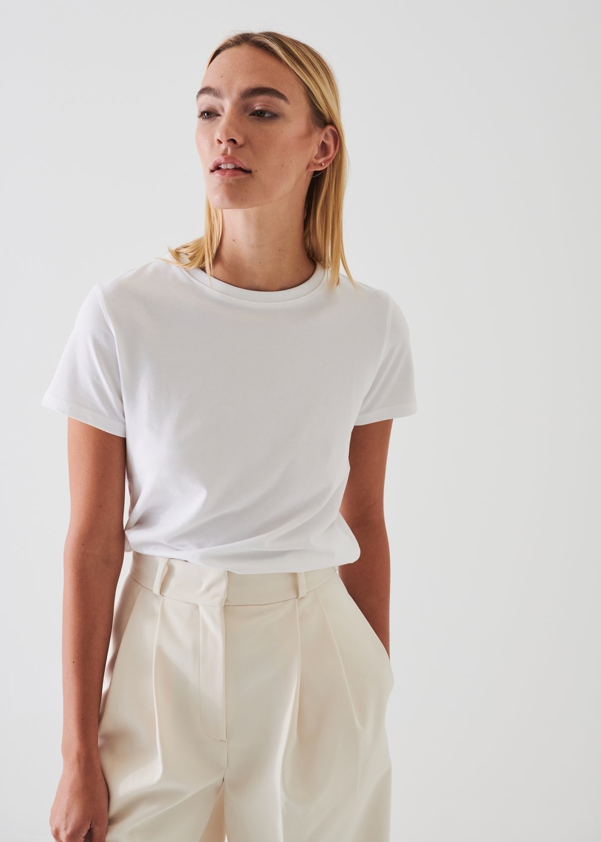 A person with long blonde hair is wearing a Patrick Assaraf SS Iconic Fitted Crew Chalk T-shirt, made of Peruvian pima cotton, tucked into high-waisted off-white trousers. The background is plain and white, and the person is looking slightly to the side.