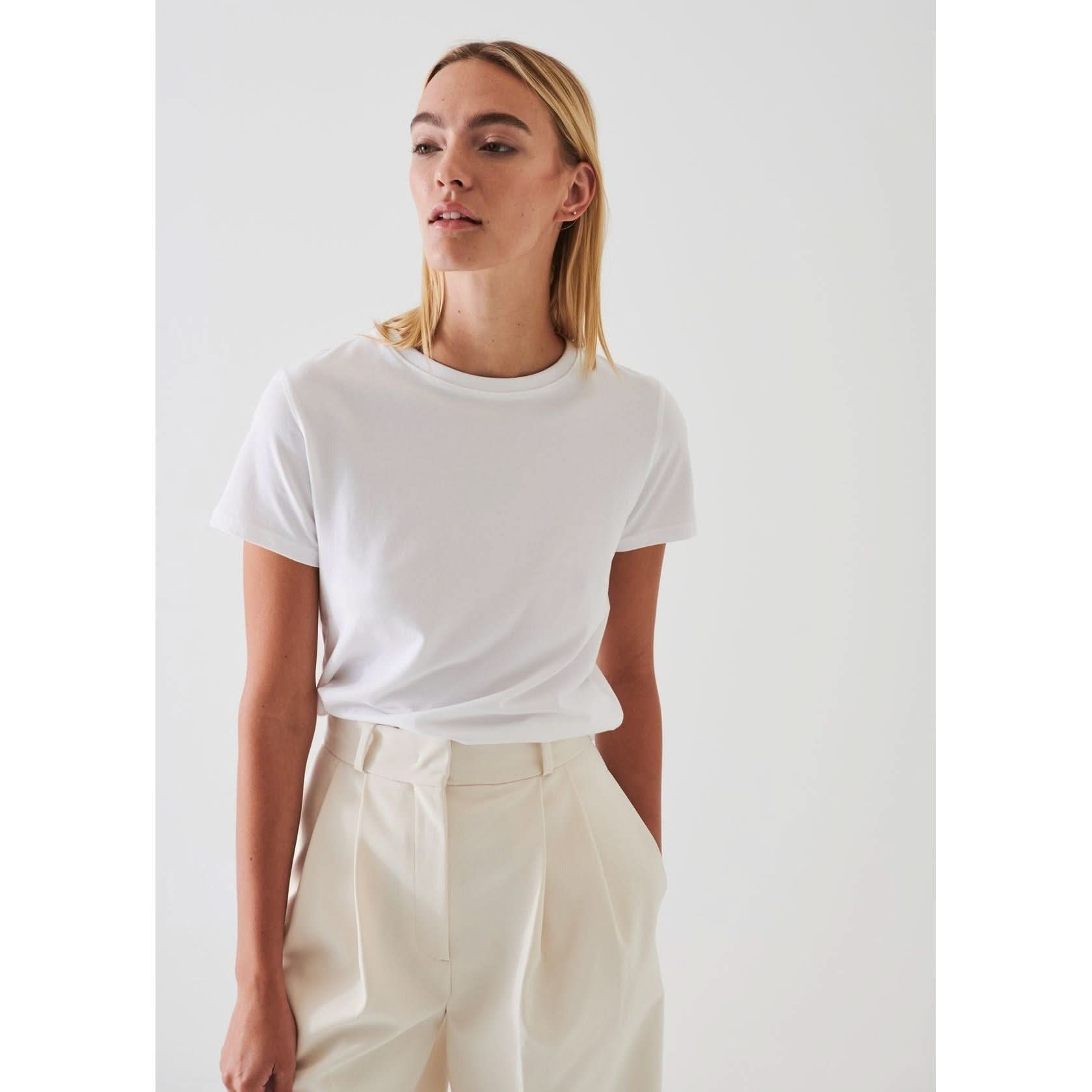 A person with long blonde hair is wearing a Patrick Assaraf SS Iconic Fitted Crew Chalk T-shirt, made of Peruvian pima cotton, tucked into high-waisted off-white trousers. The background is plain and white, and the person is looking slightly to the side.