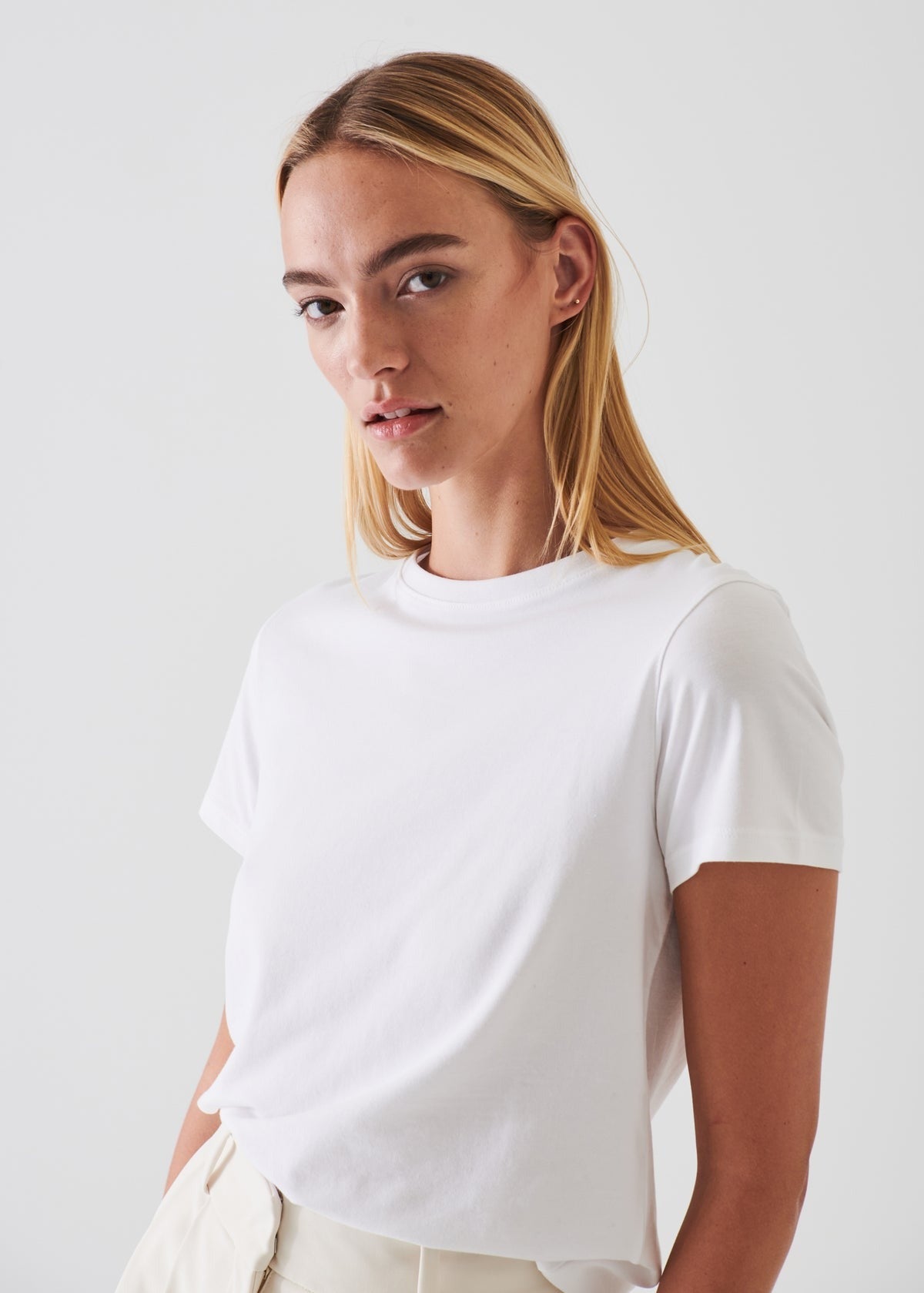 A person with long blonde hair is wearing a Patrick Assaraf SS Iconic Fitted Crew Chalk T-shirt, made from luxurious Peruvian pima cotton, along with light-colored pants. They are standing against a simple, light gray background, looking directly at the camera with a neutral expression.