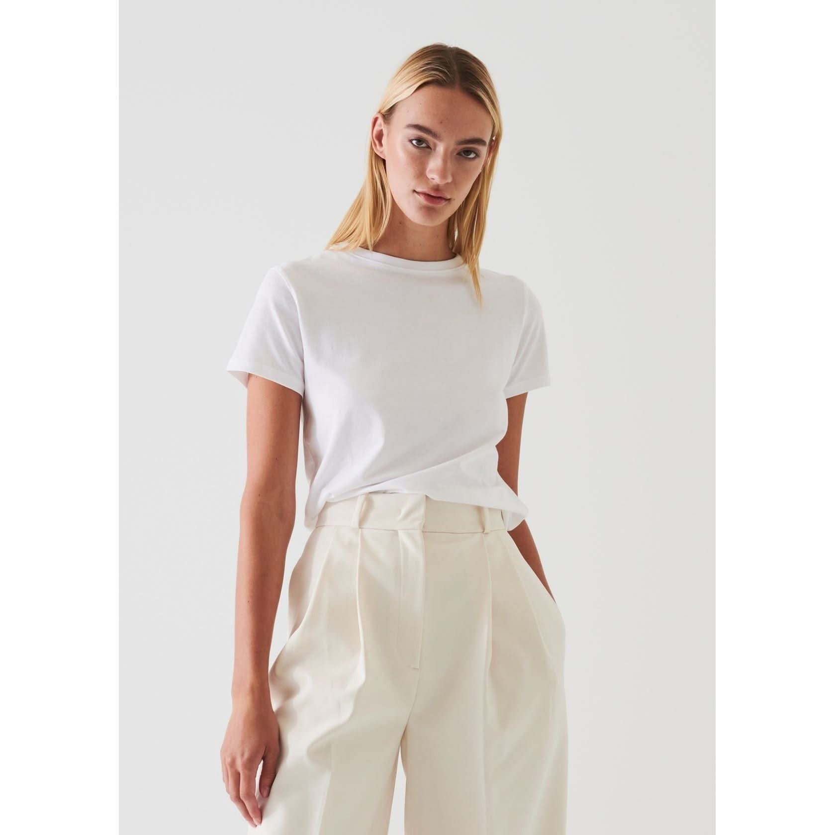 A person with straight, shoulder-length blonde hair wears the Patrick Assaraf SS Iconic Fitted Crew in a chalk color, paired with high-waisted ivory pleated trousers. They are standing against a plain white background with hands in their pockets, gazing directly at the camera.