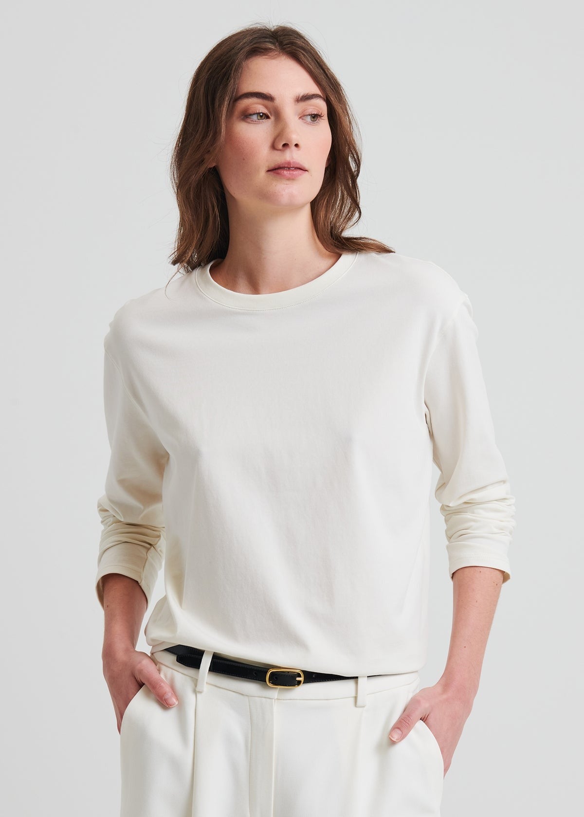 A woman with long brown hair, wearing a classic fit, Patrick Assaraf Long Sleeve Iconic Boyfriend Crew in chalk tucked into white trousers, poses with a thoughtful expression. Her hands are tucked into her pockets, and she wears a black belt. The background is plain and light-colored.