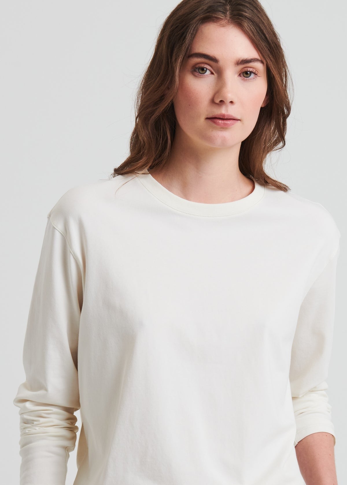 A person with long wavy hair wearing the Patrick Assaraf Long Sleeve Iconic Boyfriend Crew Chalk stands against a plain background. Their expression is neutral, and they are gazing directly at the camera.