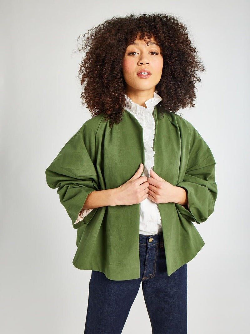 A woman with curly hair wears a Mille Amie Coat in Olive over a white ruffled blouse and dark blue jeans. She poses confidently, standing against a light grey background.