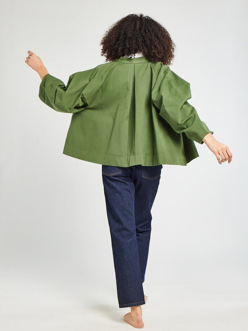 Seen from behind, a person with curly hair is wearing a Mille Amie Coat Olive featuring oversized sleeves and dark blue jeans. They are barefoot and seem to be moving forward, with one arm slightly raised against a plain, light-colored background.