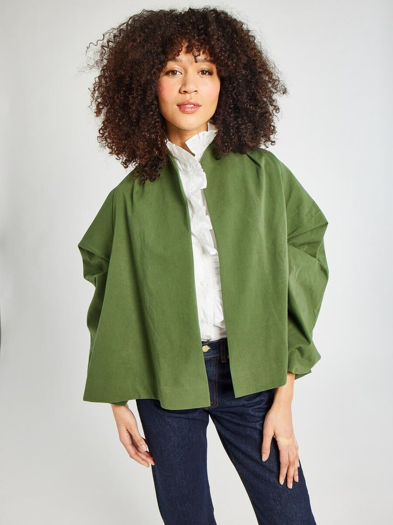 A person with curly hair is wearing the Mille Amie Coat Olive by Mille, featuring a green cape-style design with oversized sleeves. Underneath, they have on a white ruffled blouse paired with dark jeans. They are standing against a plain background and smiling softly at the camera.