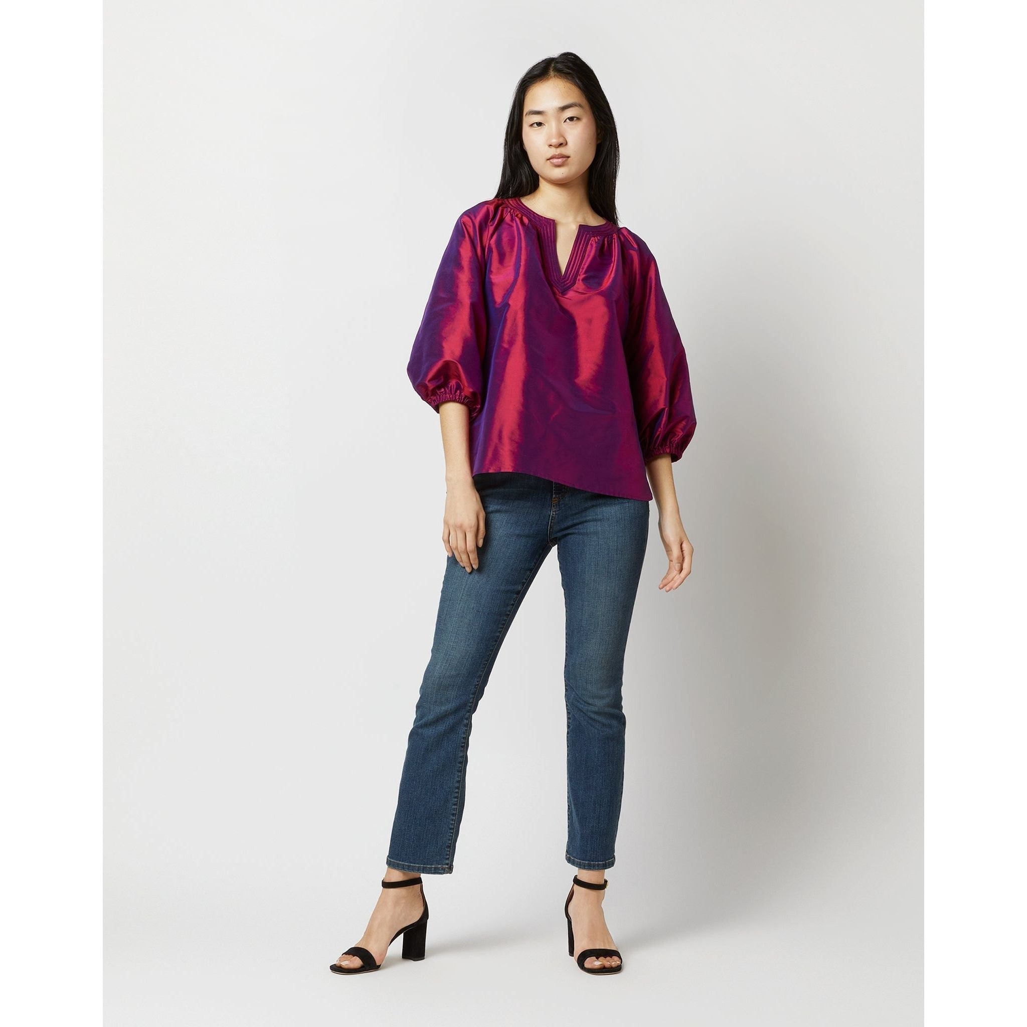 A person stands confidently against a plain backdrop, wearing the Ann Mashburn Trapunto Blouson Top in Berry Iridescent Silk Shantung, with puffed sleeves. They pair it with blue jeans and black high-heeled sandals. With long dark hair, they maintain a relaxed pose.