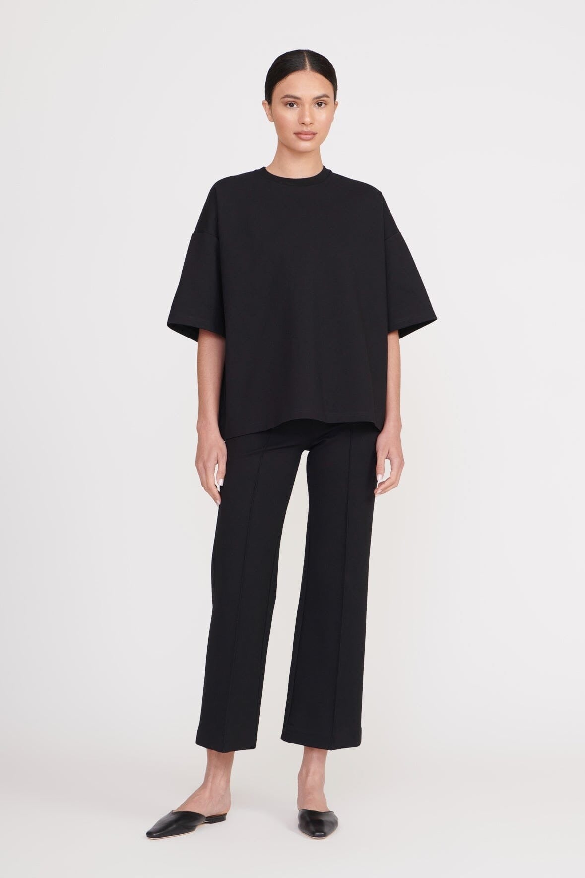 A person with dark hair tied back stands against a plain white background. They are wearing a loose-fitting black T-shirt, Staud Knack Pant in Black, and black flat shoes. With a neutral expression and hands resting by their sides, they exude a chic flair.