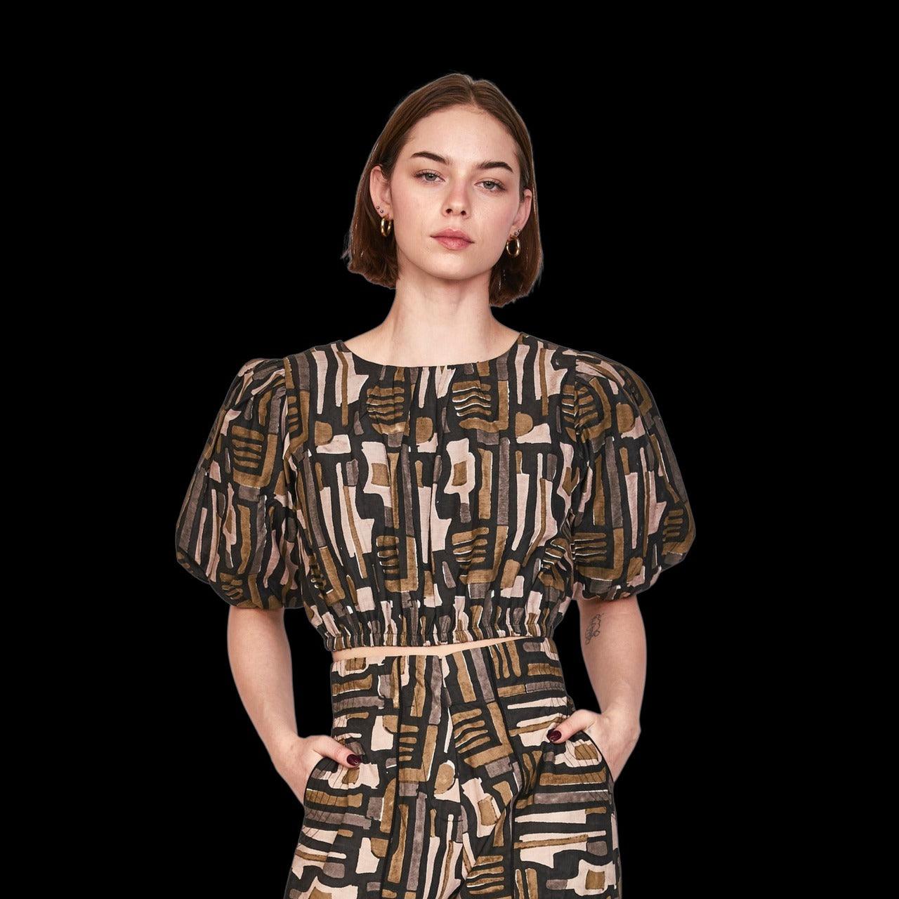 A person with short hair stands against a black background, wearing the Mirth Andaman Bubble Top in Olive Labyrinth from Mirth, which features puff sleeves and a back keyhole button closure. Their hands are in their pockets, and they have a neutral expression on their face. They are accessorized with small hoop earrings.