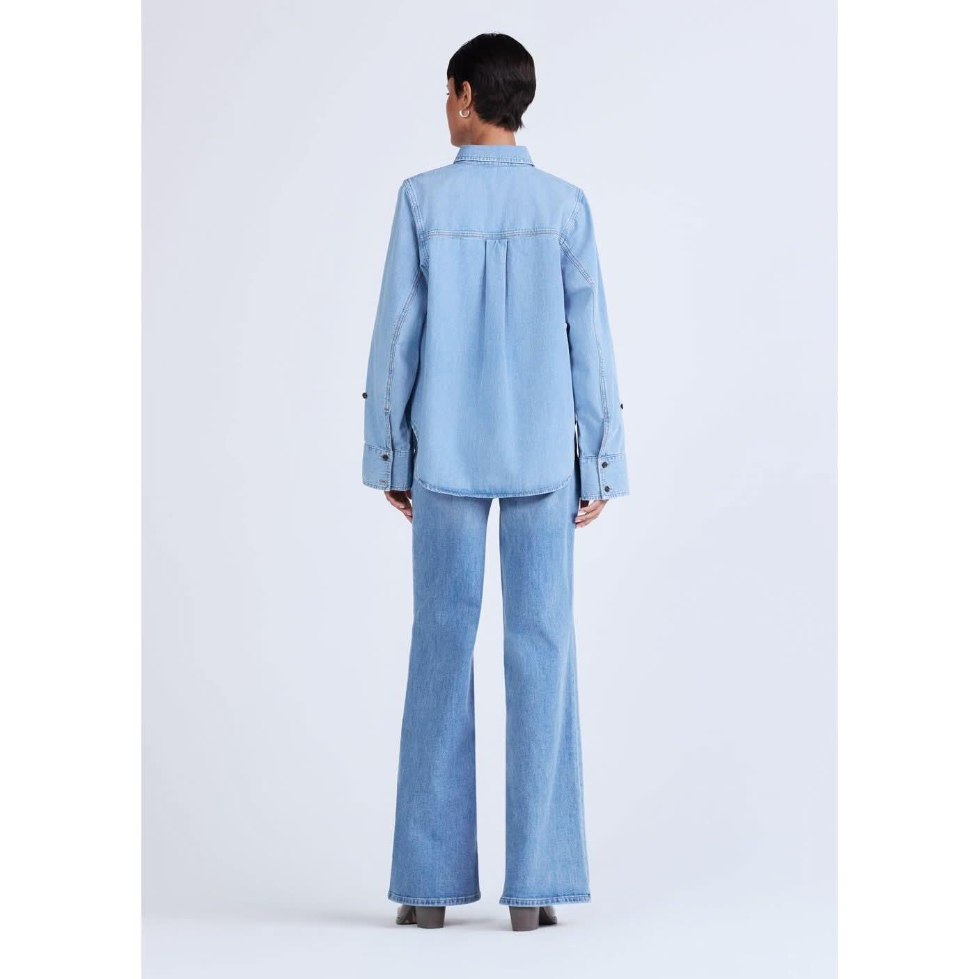 A person with short, dark hair is standing with their back to the camera, wearing an ultra soft Derek Lam 10 Crosby Wesley Denim Shirt featuring button sleeve detail and matching wide-leg jeans. The outfit has a casual yet stylish look against a plain off-white background.