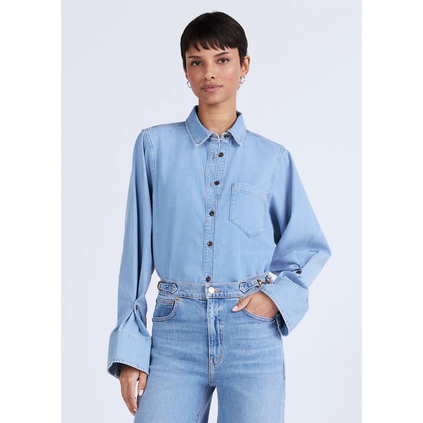 A person with short dark hair is posing against a plain background, wearing a Derek Lam 10 Crosby Wesley Denim Shirt in Dover Light with large cuffs and metal buttons, tucked into high-waisted denim jeans. They have a neutral expression and are standing with one hand in a pocket.
