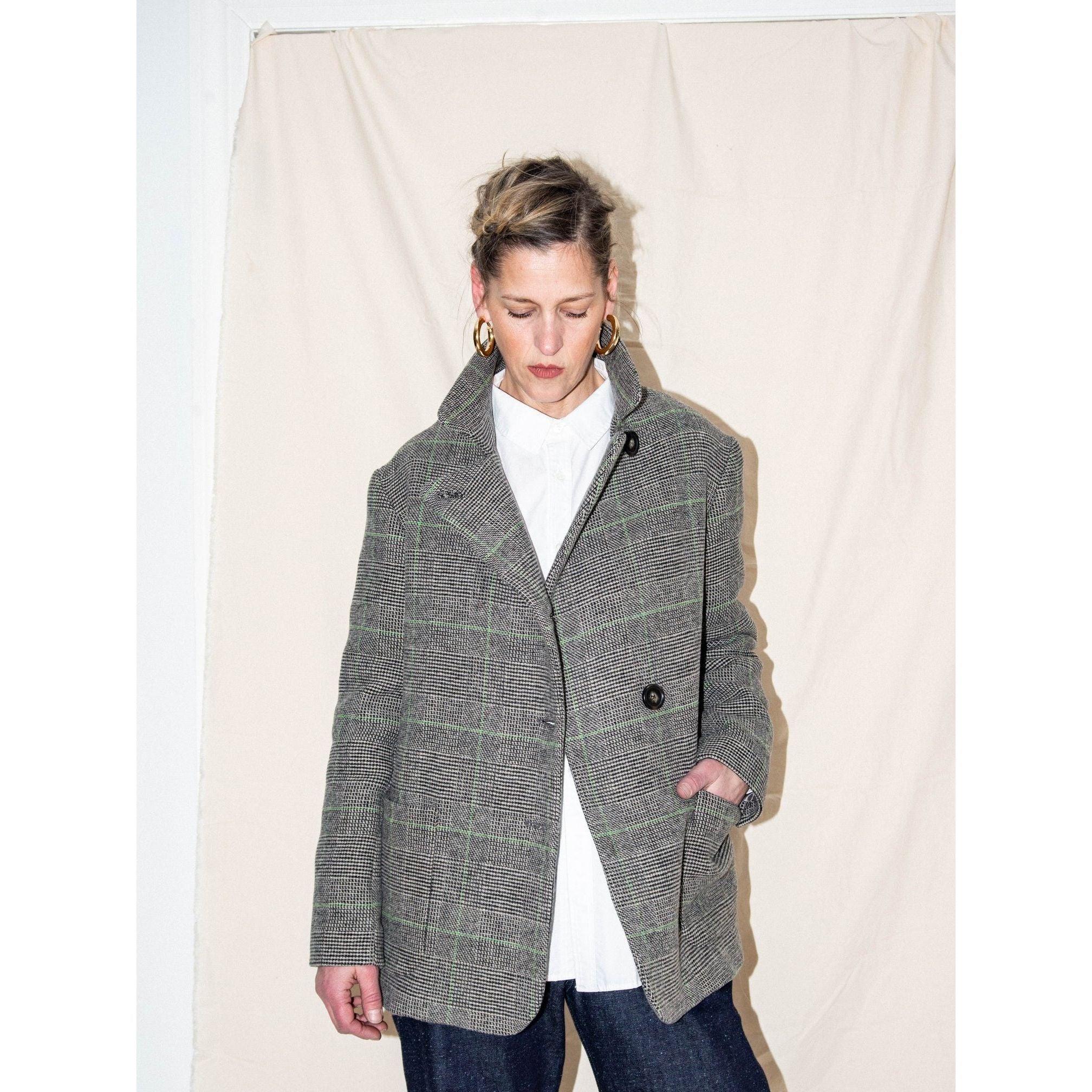 A person with short hair stands in front of a beige backdrop, wearing the Heather Harlan Grandad Blazer Jacket in plaid along with dark pants. Their white shirt is crisp beneath the ensemble, which is accented by large hoop earrings. With eyes gently closed, they embody the elegance of sustainable fashion.