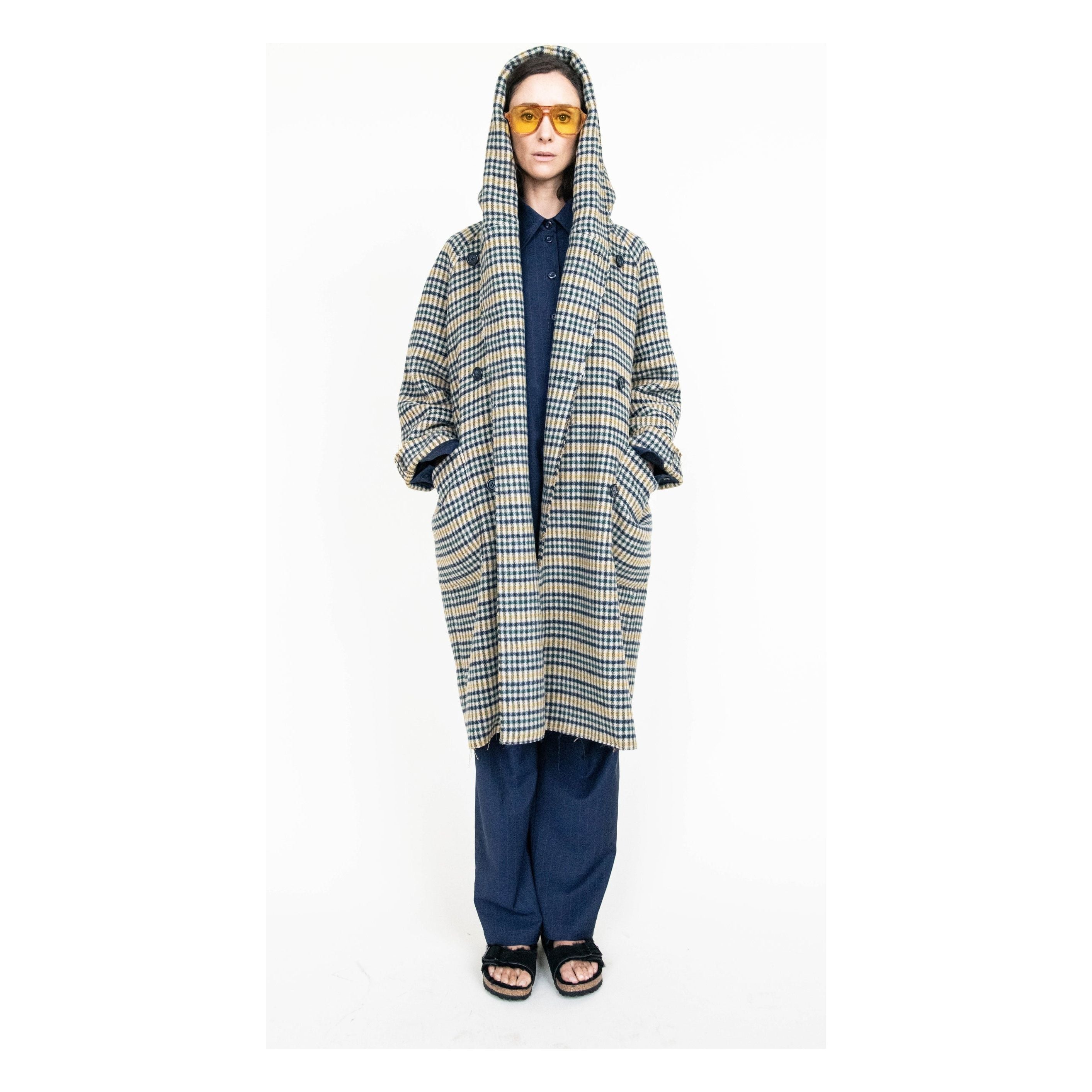 A person wears a Heather Harlan Hooded Blanket Coat, crafted from recycled Italian wool, against a plain backdrop. Their blue and beige plaid coat is distinguished by an oversized draped hood. Completing the chic outfit are dark pants and yellow sunglasses, with hands tucked in pockets and feet in sandals.