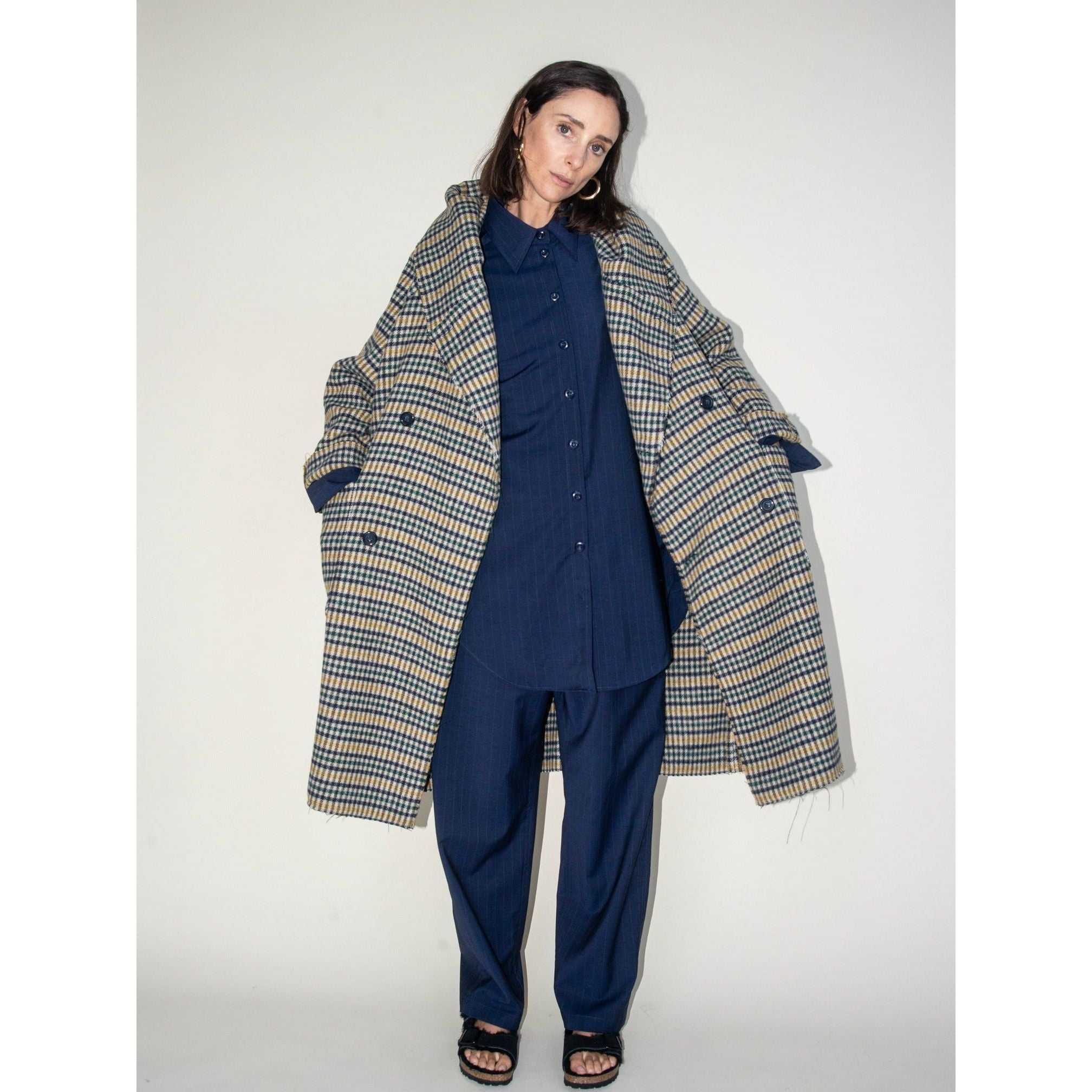 In front of a plain backdrop, a person showcases the Heather Harlan Hooded Blanket Coat, an oversized plaid piece crafted from recycled Italian wool. They pair it effortlessly over a navy blue shirt and pants and match it with black sandals. With long dark hair cascading down, they gaze directly at the camera.