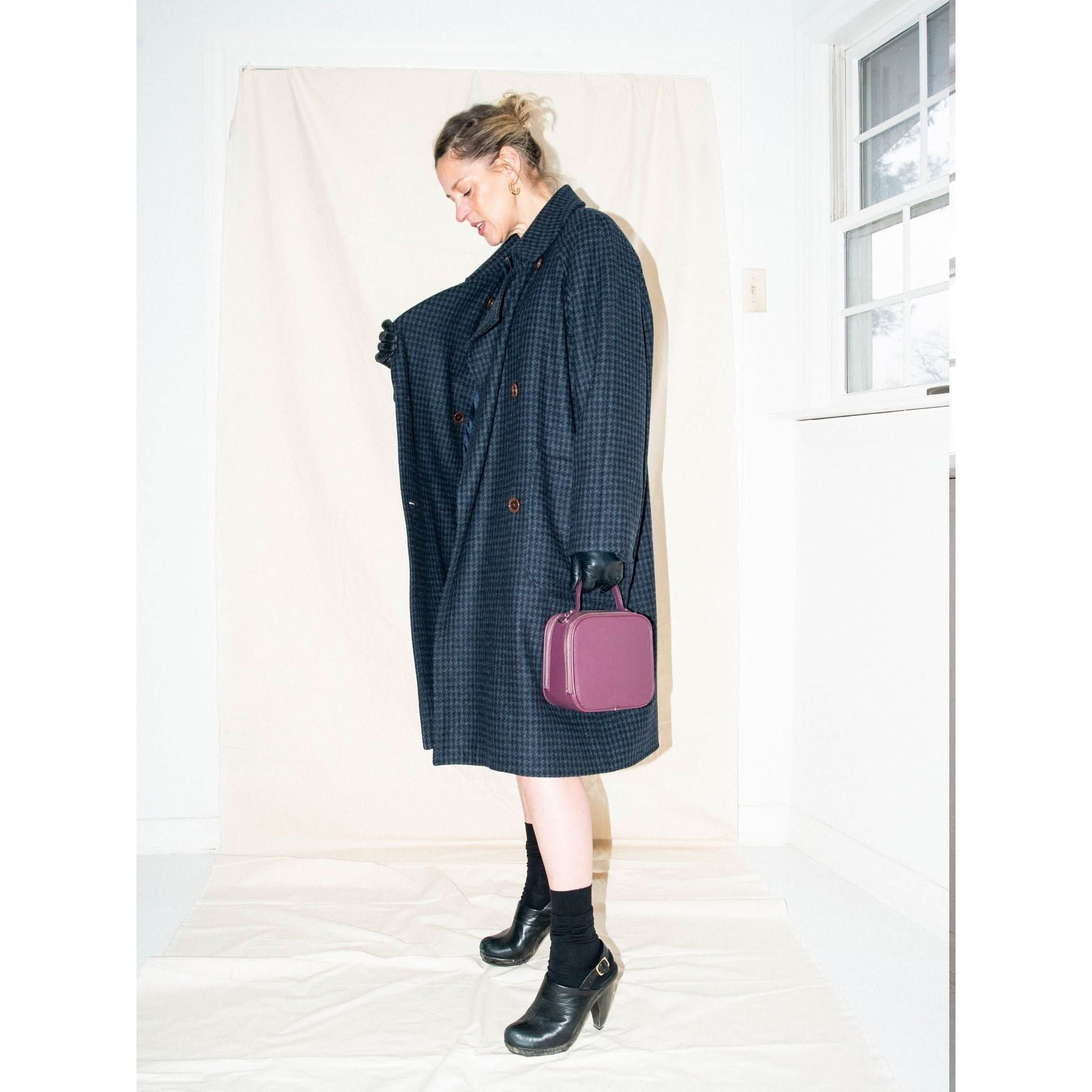 A person in a long, dark Heather Harlan Houndstooth Overcoat stands holding a small purple bag. They wear black gloves, socks, and heeled shoes. The scene is set against a simple, light backdrop with a window to the side.