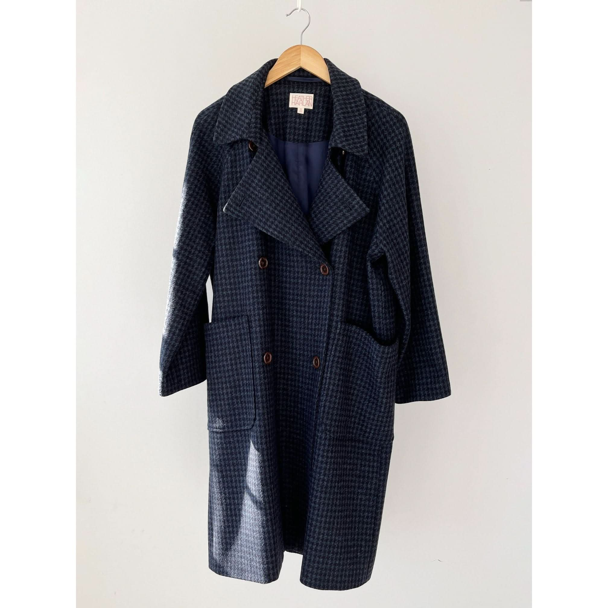 The Heather Harlan Houndstooth Overcoat, a long, dark navy piece crafted from recycled wool-cashmere, showcases a subtle checkered pattern along with double-breasted buttons, oversized pockets, and a wide collar. It is displayed on a wooden hanger against a plain light background.