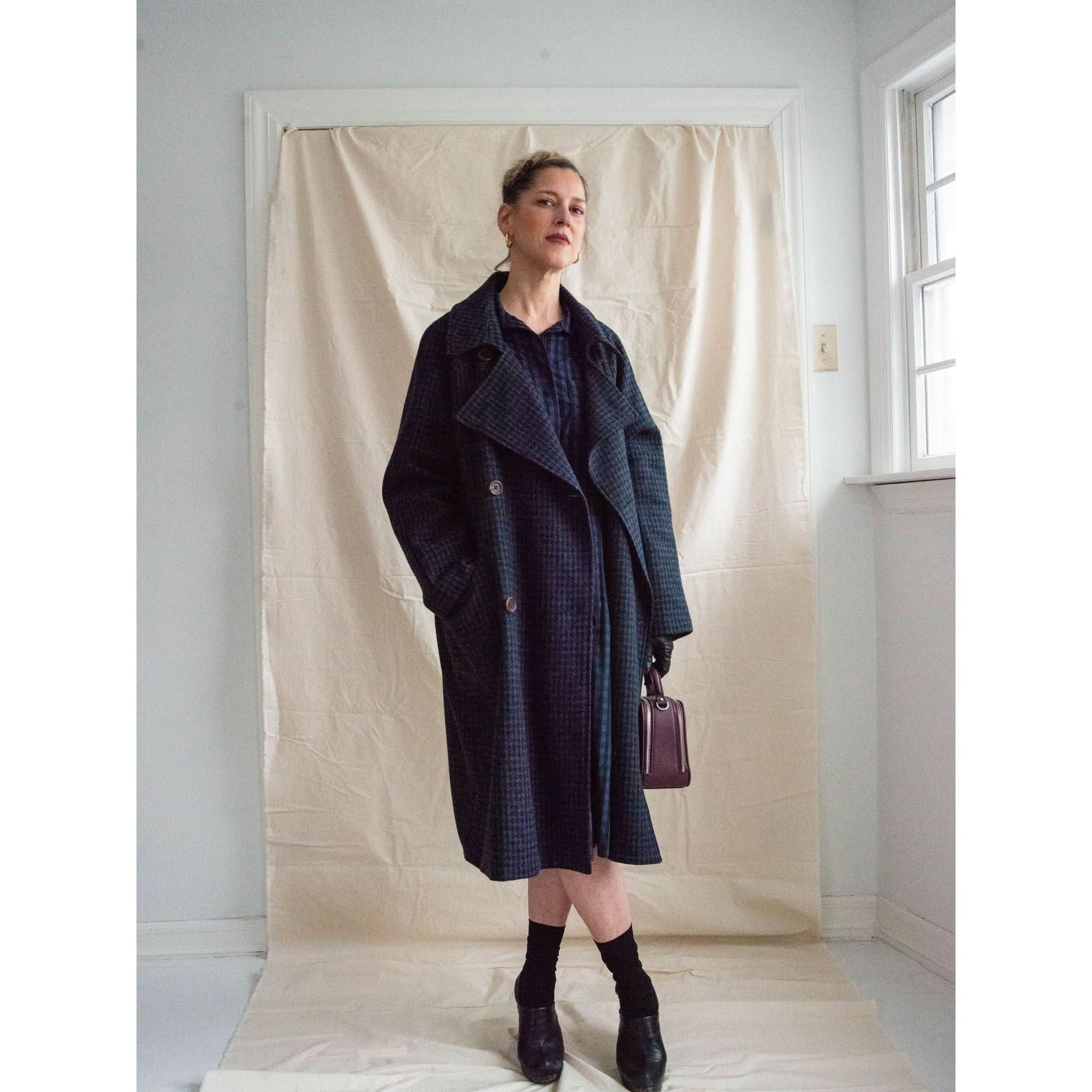 In a softly lit minimalist indoor setting, a person stands against a beige backdrop, wearing the Heather Harlan Houndstooth Overcoat featuring oversized pockets. They have short hair and carry a small purple bag. Natural light from a nearby window gently illuminates the elegance of the recycled wool-cashmere fabric.