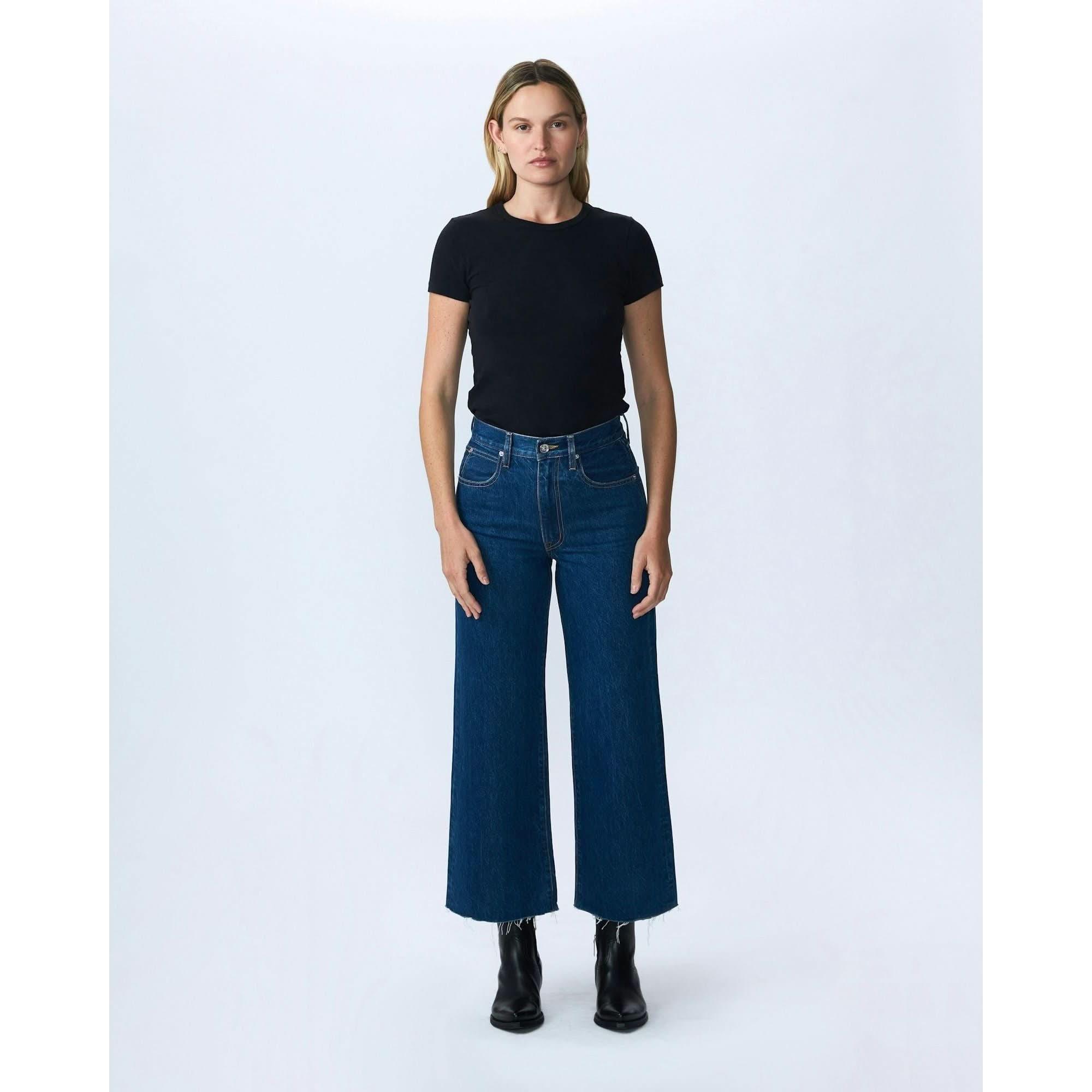 A person stands against a plain background wearing a black t-shirt, Slvrlake Denim Grace Crop Revolution high-rise wide-leg jeans from SLVRLAKE in the color Revolution, and black ankle boots. They have shoulder-length hair and a neutral expression.