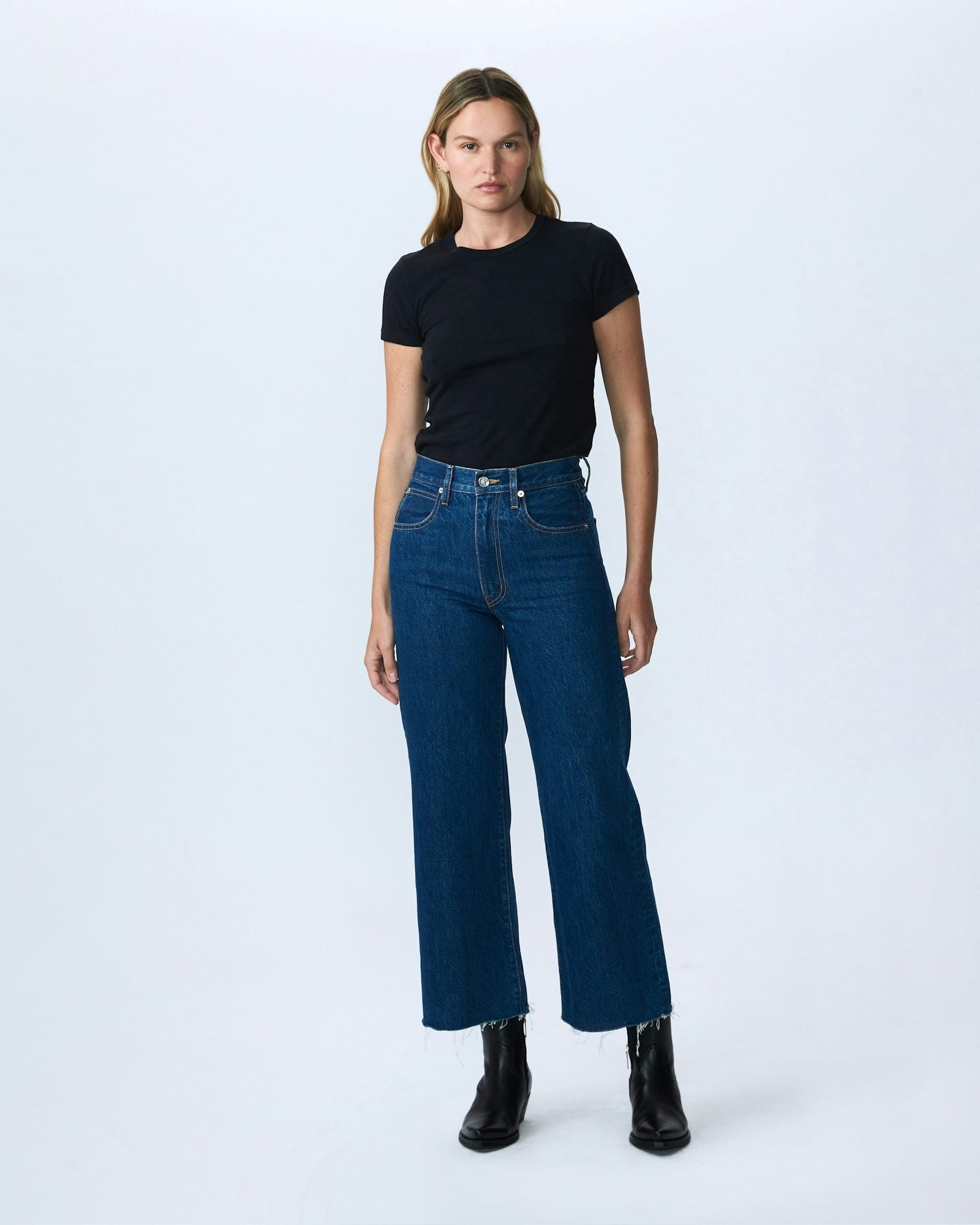 A person with shoulder-length hair stands against a plain white background. They are wearing a black t-shirt tucked into SLVRLAKE's Slvrlake Denim Grace Crop Revolution high-rise, dark blue jeans with a cropped wide leg that stops above the ankles. They are also wearing black ankle boots.