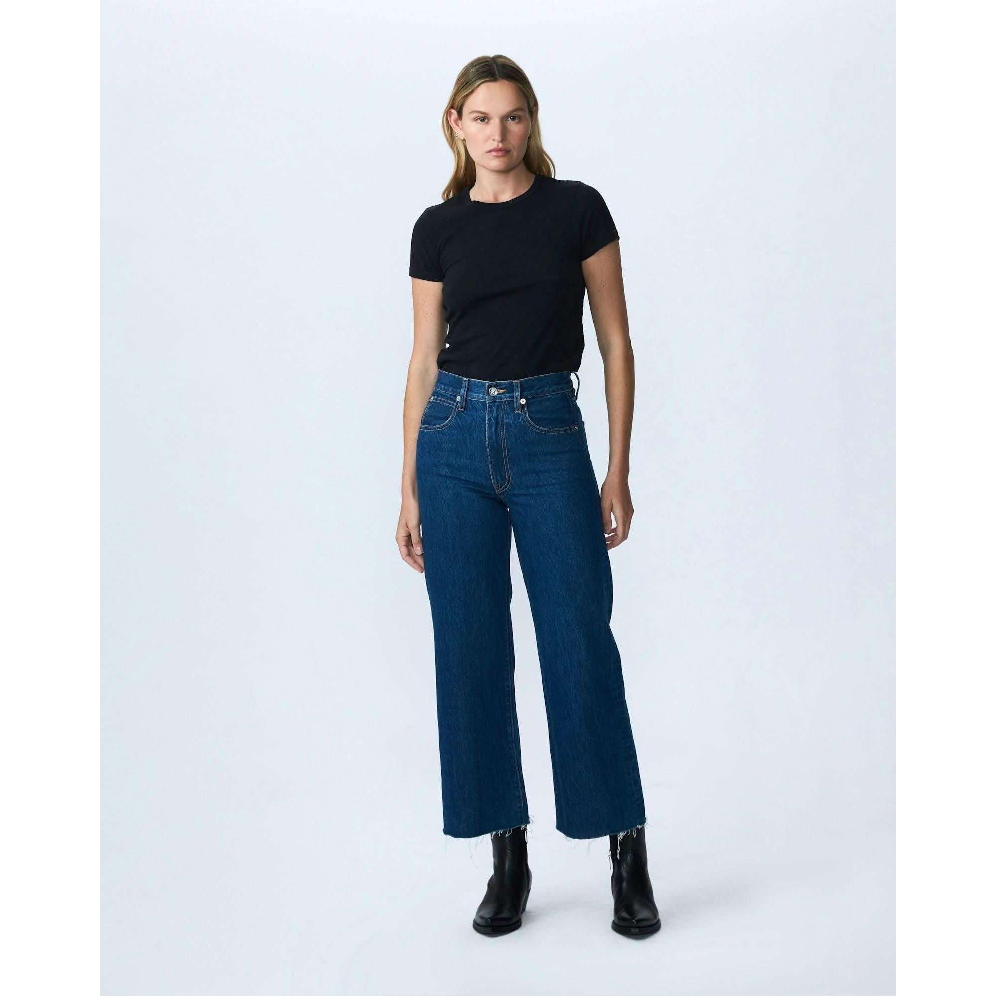 A person with shoulder-length hair stands against a plain white background. They are wearing a black t-shirt tucked into SLVRLAKE's Slvrlake Denim Grace Crop Revolution high-rise, dark blue jeans with a cropped wide leg that stops above the ankles. They are also wearing black ankle boots.