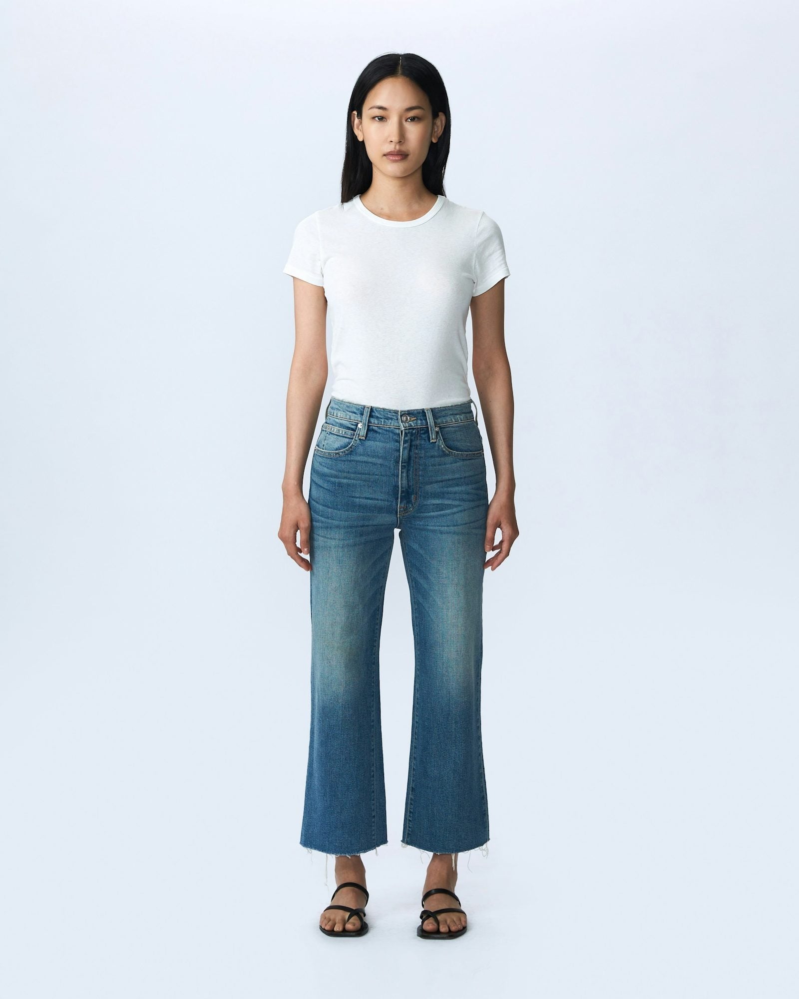 A woman stands against a plain light-colored background. She is wearing a fitted white T-shirt, SLVRLAKE's Grace Crop Blue Bird high-rise blue jeans with a slight flare and raw hem, and black sandals. She has long straight hair and a neutral expression, with her arms relaxed by her sides. The look reflects effortless style made in the U.S.A.