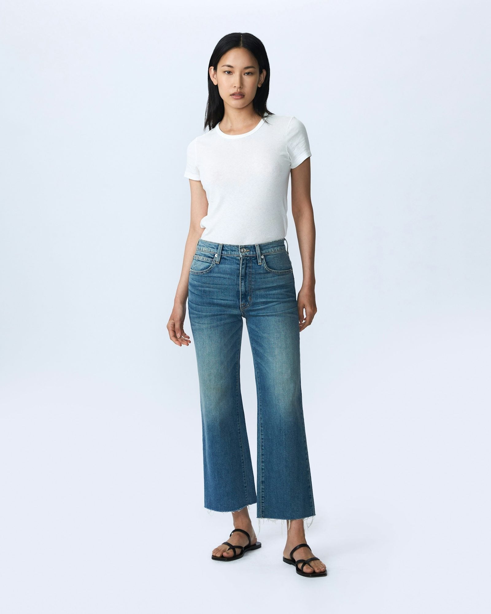 A person with long hair stands against a plain background, wearing a white short-sleeve T-shirt, Slvrlake Denim Grace Crop Blue Bird high-rise jeans with wide legs by SLVRLAKE, and black strappy sandals. The person has a relaxed pose with arms resting by their sides.