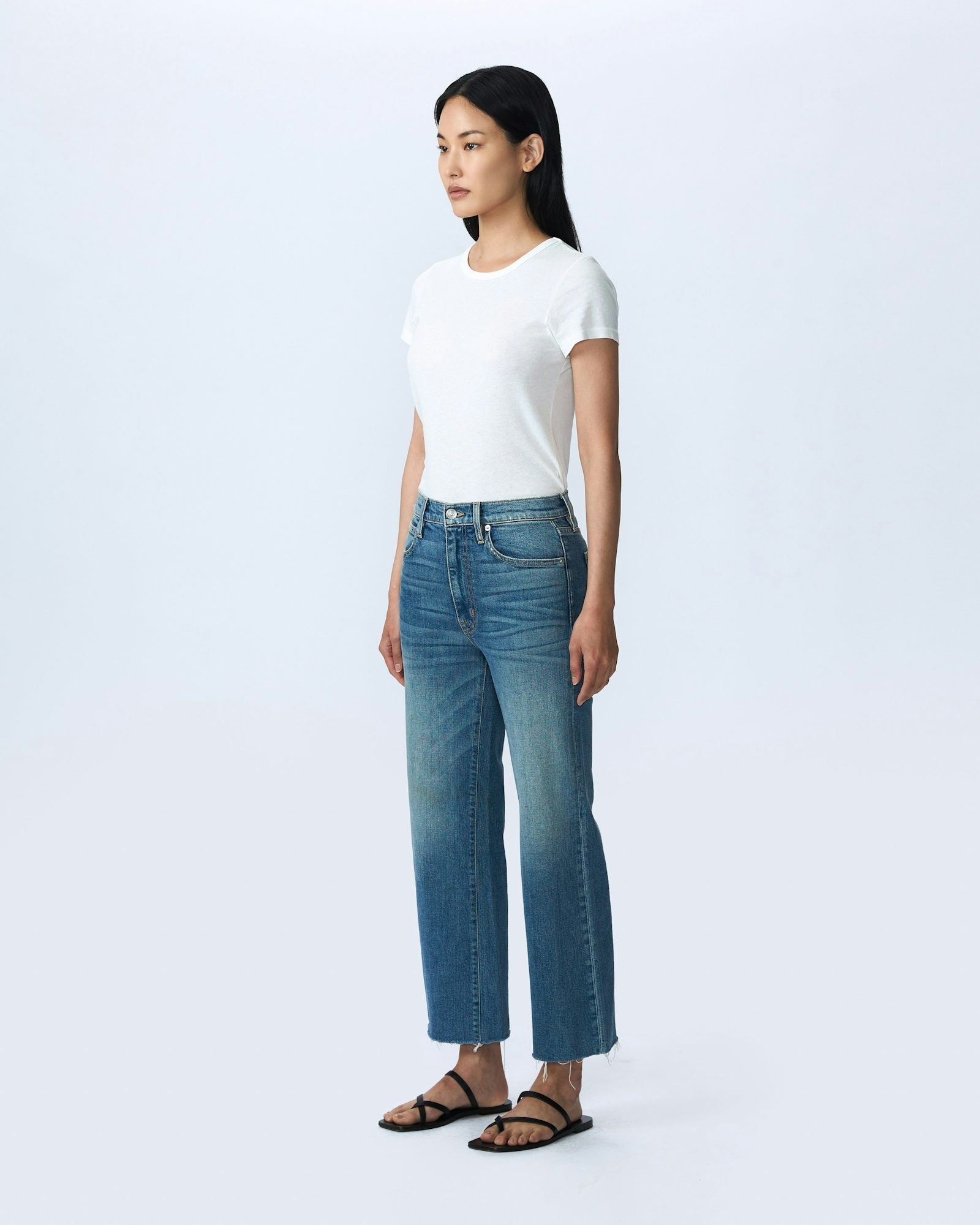 A person stands against a plain white background, wearing a fitted white t-shirt, high-waisted Slvrlake Denim Grace Crop Blue Bird jeans from SLVRLAKE with a slight flare at the hem, and black sandals. The high-rise jeans are proudly Made In U.S.A. The person has long, straight, dark hair and is standing confidently with hands by their sides.