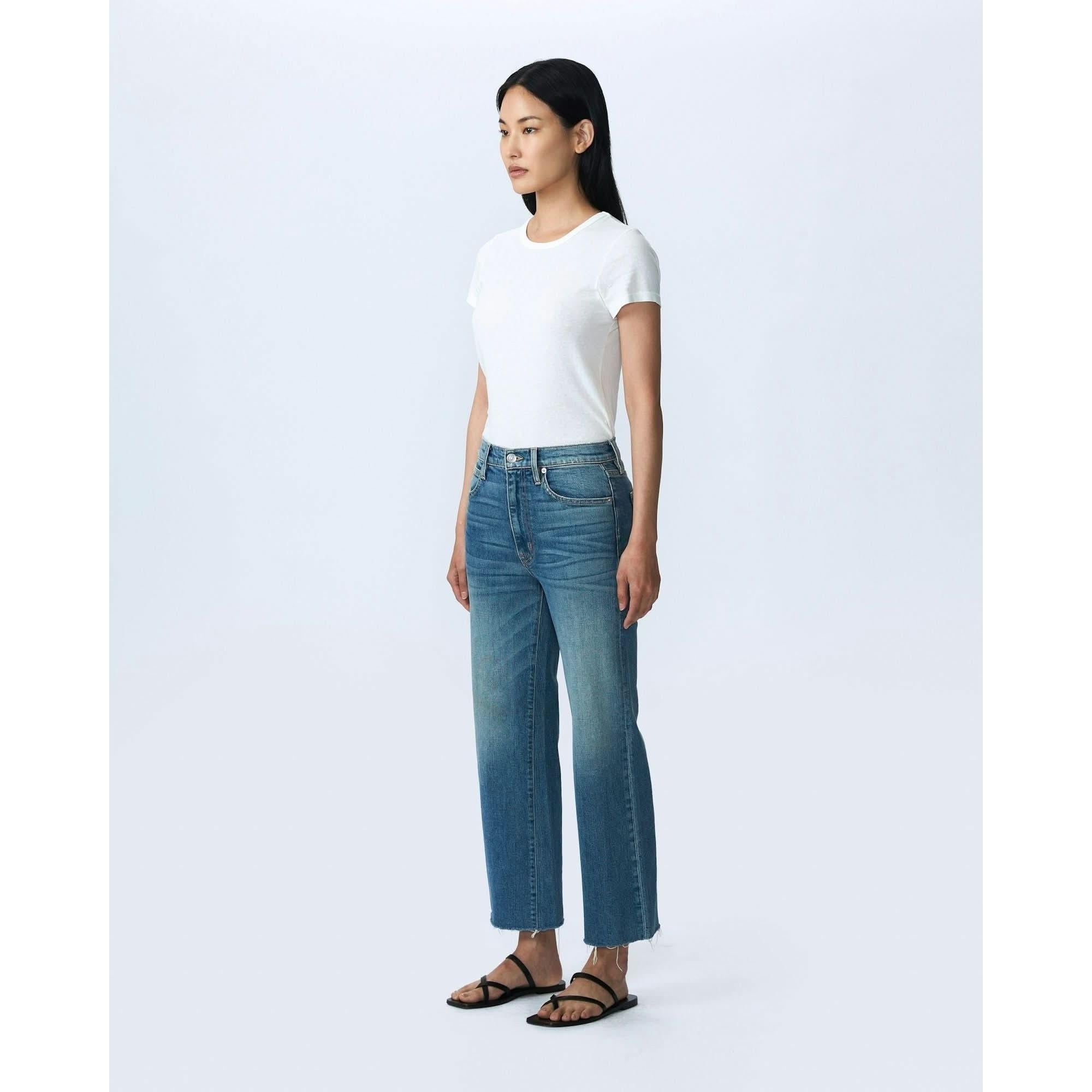 A person stands against a plain white background, wearing a fitted white t-shirt, high-waisted Slvrlake Denim Grace Crop Blue Bird jeans from SLVRLAKE with a slight flare at the hem, and black sandals. The high-rise jeans are proudly Made In U.S.A. The person has long, straight, dark hair and is standing confidently with hands by their sides.