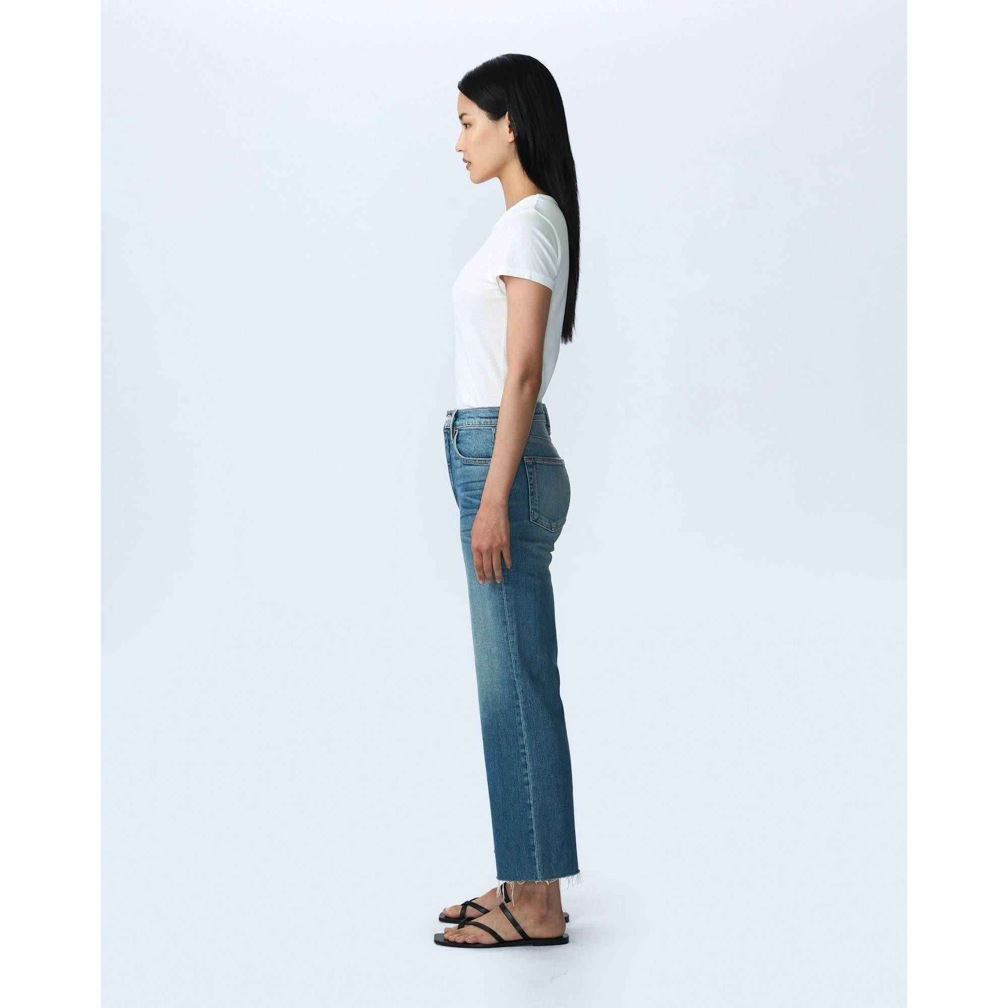 A person with long dark hair stands in profile against a plain white background. They are wearing a white short-sleeved t-shirt, SLVRLAKE denim Grace Crop Blue Bird high-rise jeans with wide legs, and black sandals. The person is looking straight ahead with their arms relaxed at their sides.