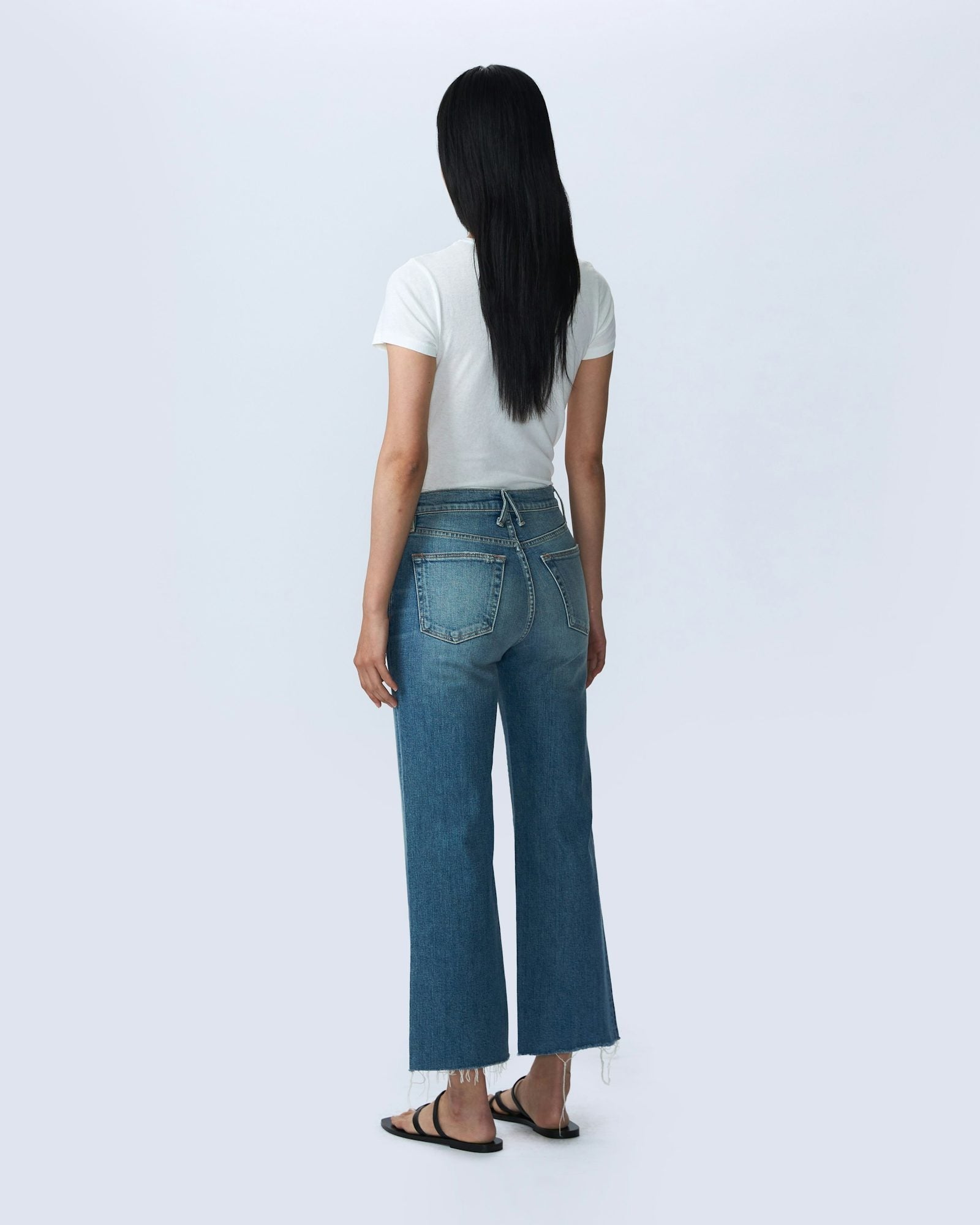 A person with long black hair stands facing away, wearing a white short-sleeve T-shirt and SLVRLAKE's Grace Crop Blue Bird high-rise, wide-leg jeans. They are also wearing black sandals. The background is a plain light grey color.