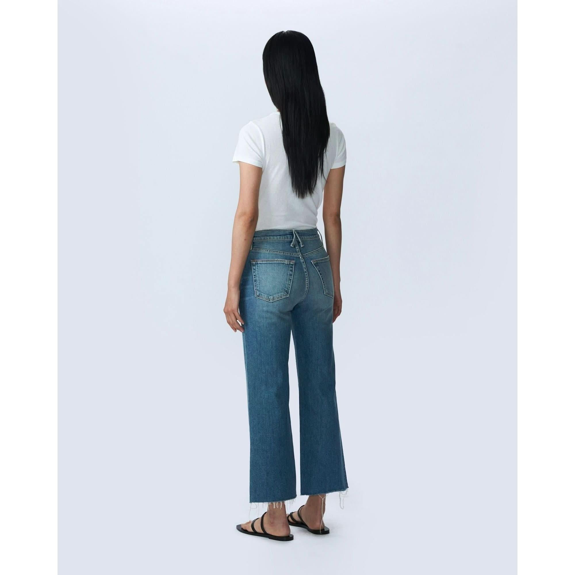 A person with long black hair stands facing away, wearing a white short-sleeve T-shirt and SLVRLAKE's Grace Crop Blue Bird high-rise, wide-leg jeans. They are also wearing black sandals. The background is a plain light grey color.