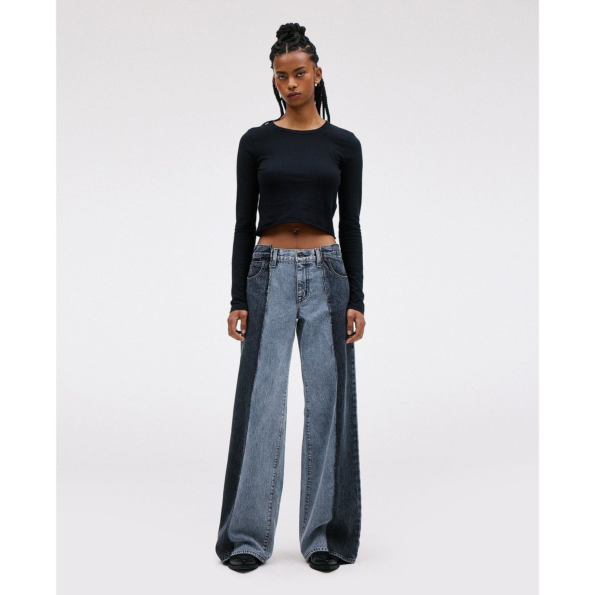 A person with long braided hair stands confidently, wearing the SLVRLAKE RE-Work Mica Panel Lunar Eclipse black long-sleeve crop top along with high-rise cropped wide-leg blue jeans. The neutral background highlights their crafted outfit and posture.