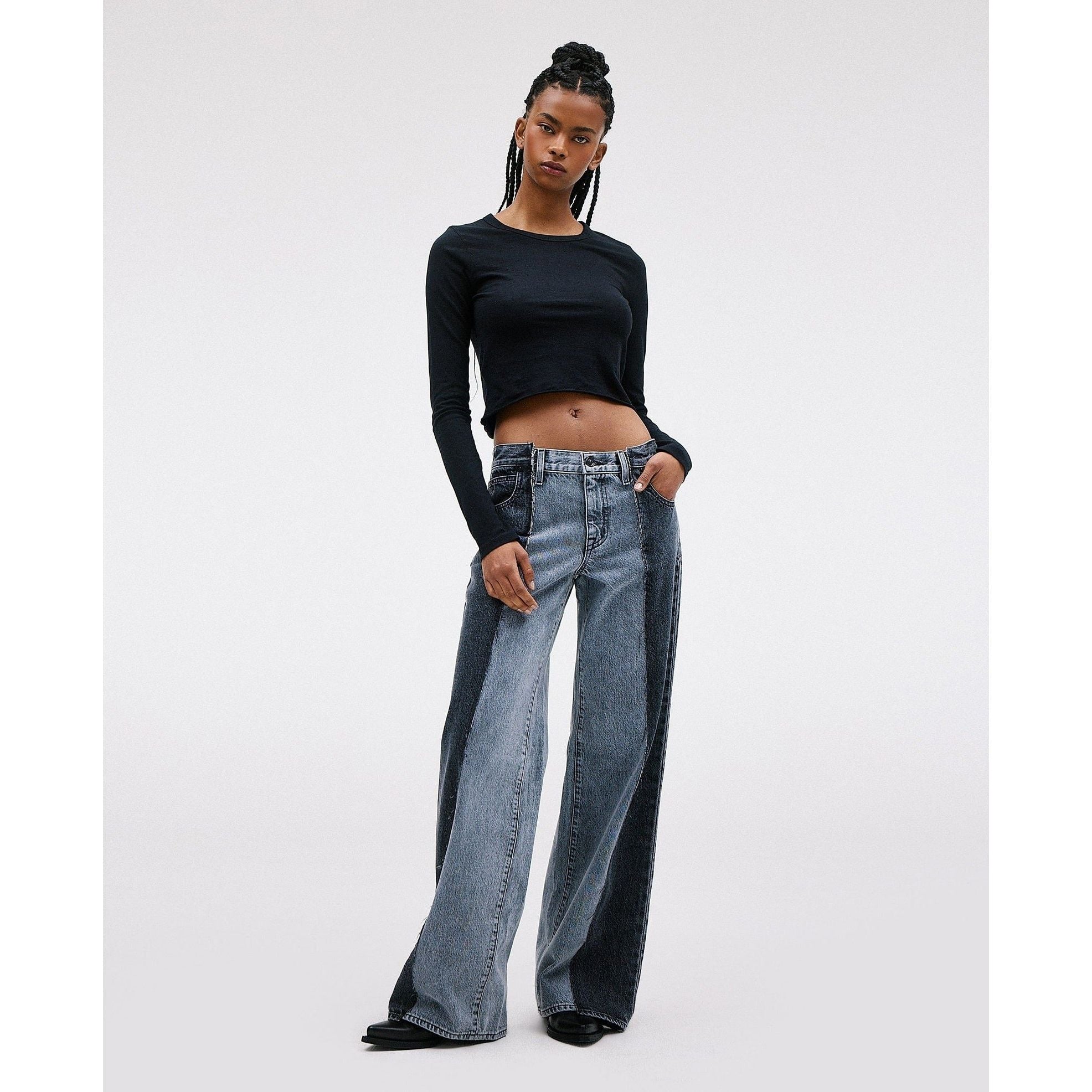 A person with long braided hair stands confidently in the SLVRLAKE RE-Work Mica Panel Lunar Eclipse, featuring a black long-sleeve crop top and high-rise, cropped wide-leg jeans. Their left hand is casually placed in a pocket, posing against a plain light background.