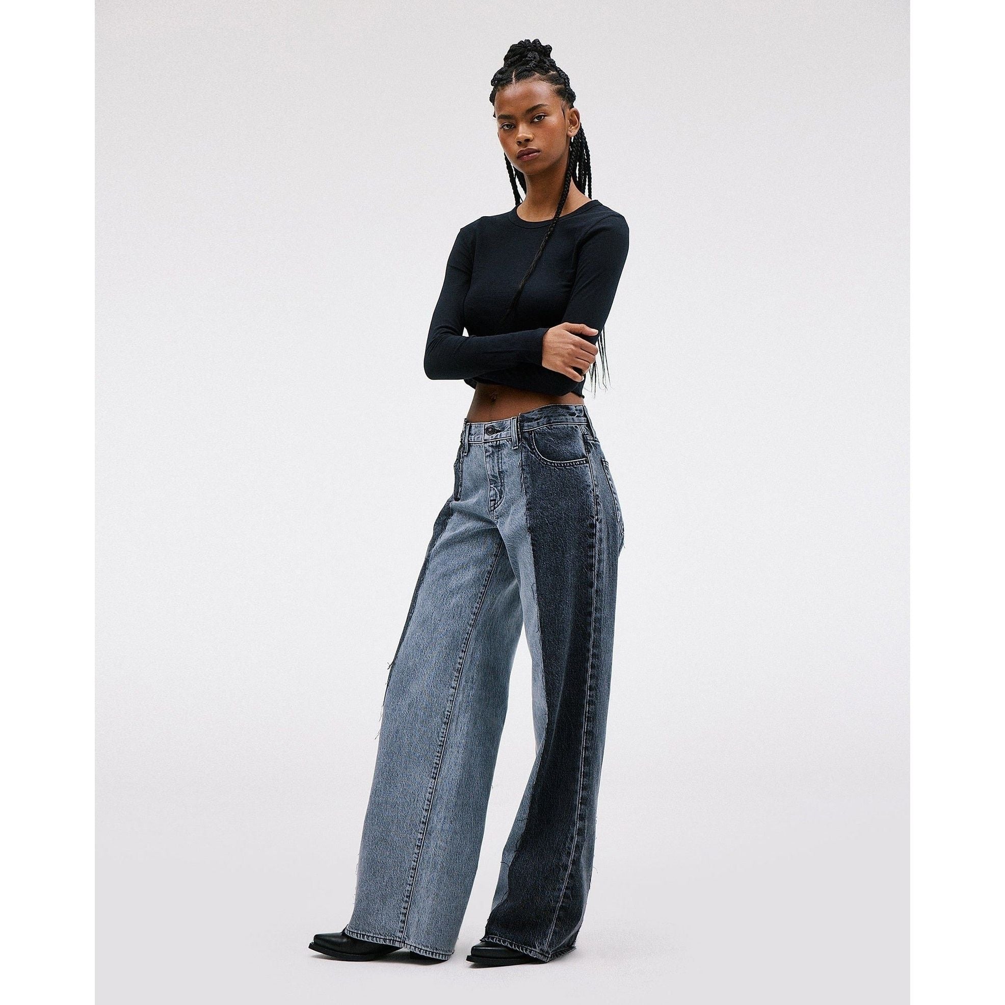 A person with long braided hair is wearing the Slvrlake RE-Work Mica Panel Lunar Eclipse, a black long-sleeve top and high-rise wide-leg jeans by SLVRLAKE featuring a crafted patchwork design. They stand against a plain white background, one arm resting on their hip.