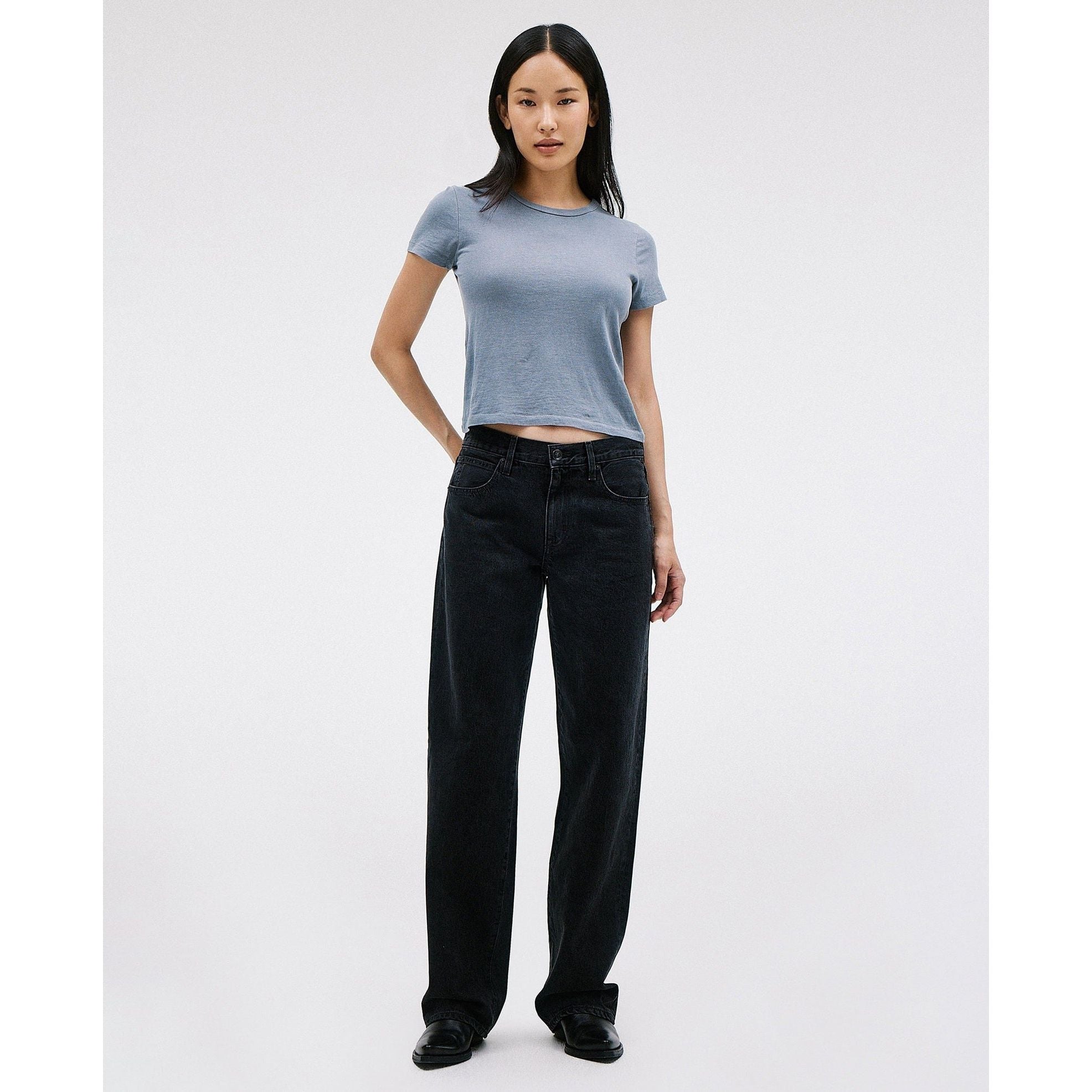 A person with long black hair stands against a plain background, wearing the Slvrlake Tess Long Shadow Ridge from SLVRLAKE—a light gray cropped T-shirt—paired with low-rise black wide-leg jeans made from 100% cotton and sleek black shoes. The pose is relaxed and confident.