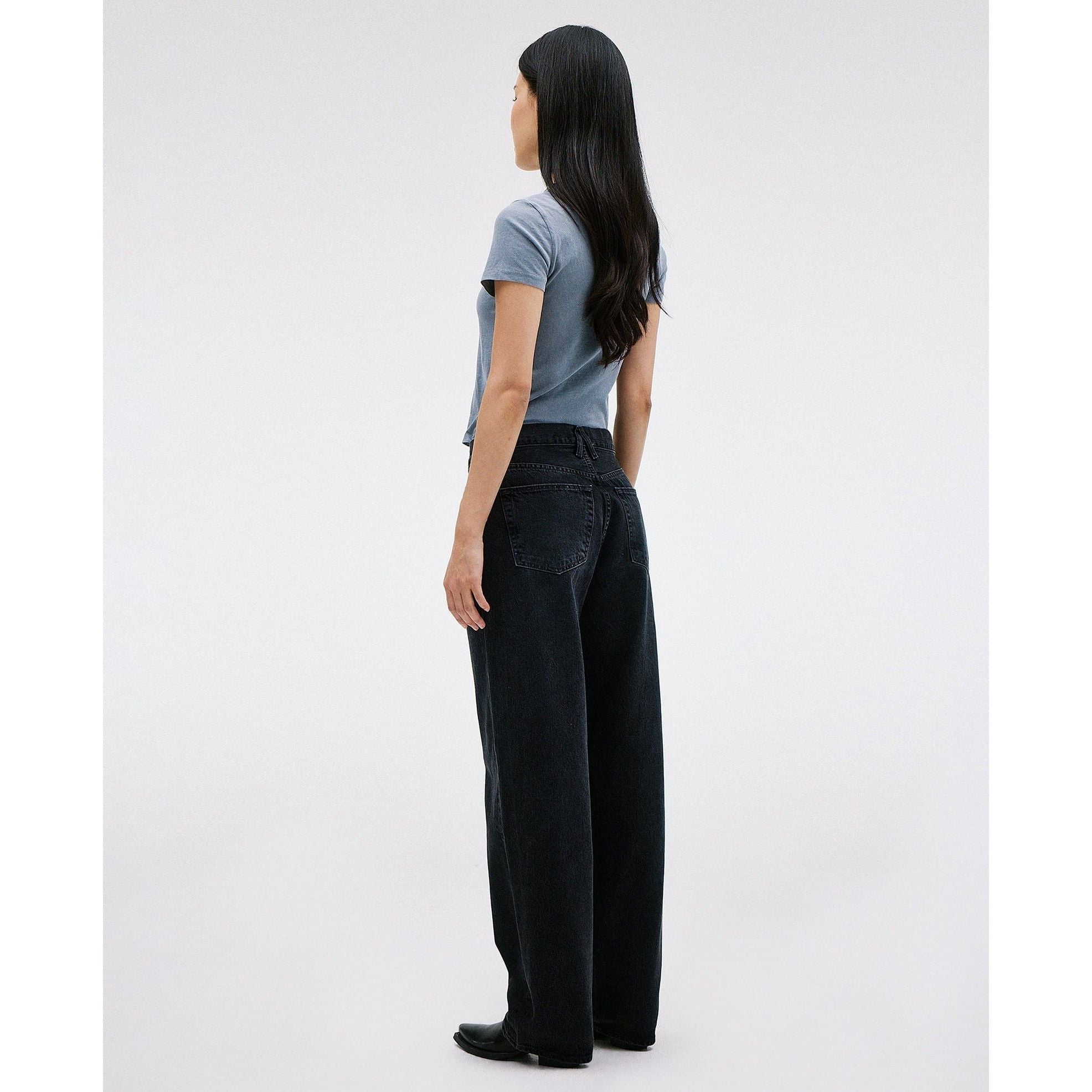 A person with long black hair stands sideways in a light blue t-shirt and the Slvrlake Tess Long Shadow Ridge wide-leg, low-rise black jeans from SLVRLAKE, made of 100% cotton. The background is plain white.