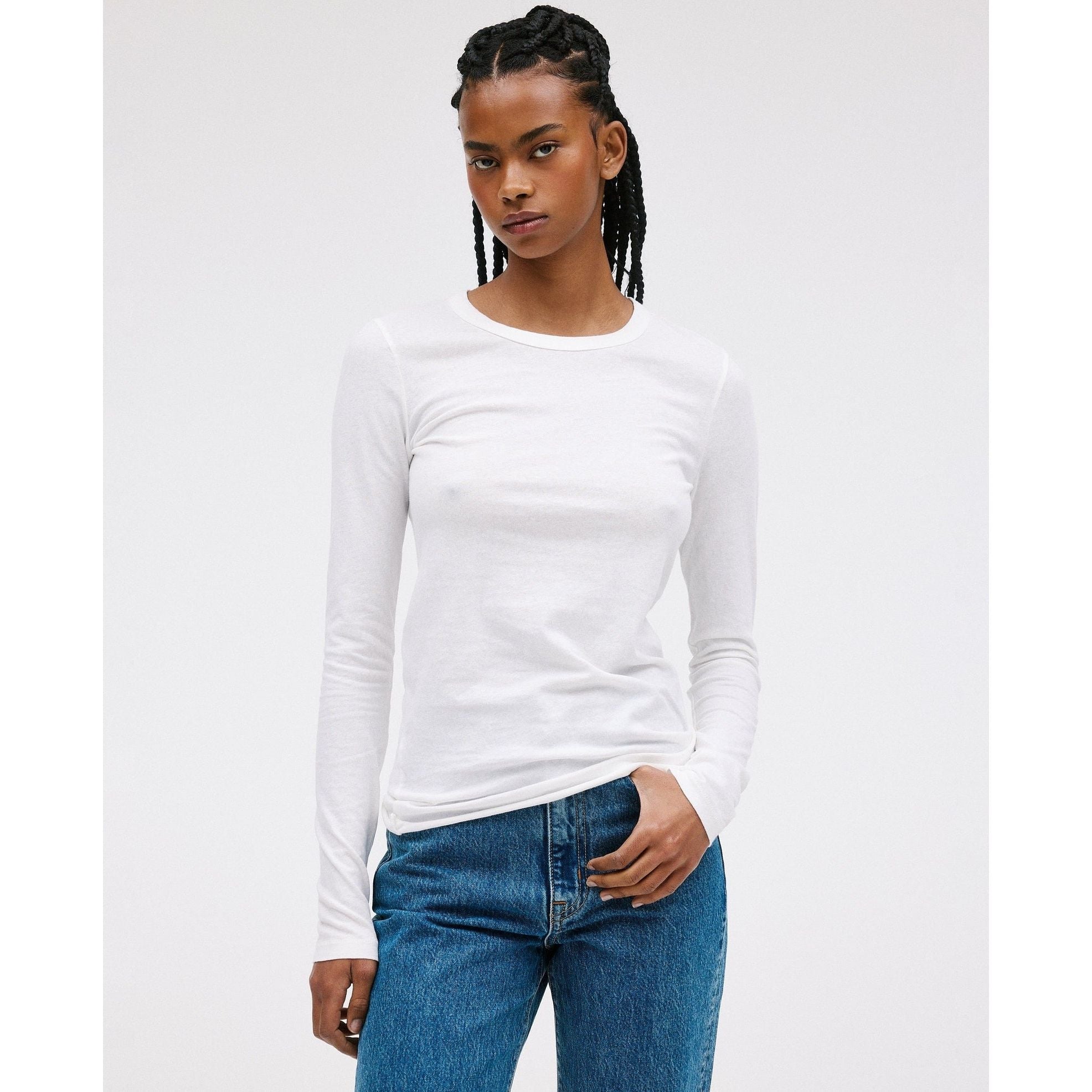 A person stands against a plain background wearing the SLVRLAKE Simple Long Sleeve Tee in Natural White, made of 100% cotton, paired with blue, wide-leg jeans. Their hair is braided, and they are looking directly at the camera.