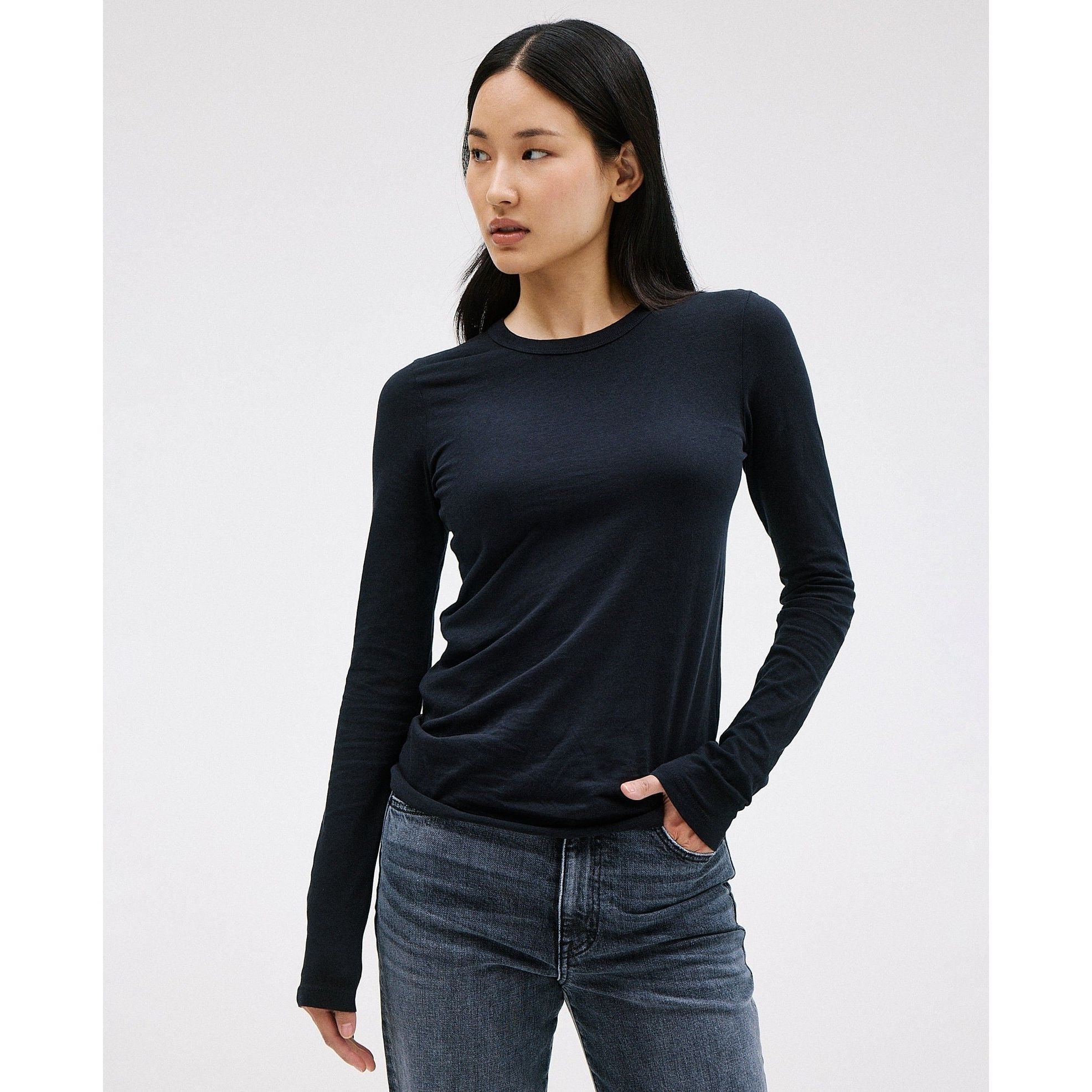 A person with long black hair stands against a plain background, wearing the SLVRLAKE Simple Long Sleeve Tee in Raven and jeans. They look to the side with one hand in their pocket, exuding a casual yet stylish vibe.