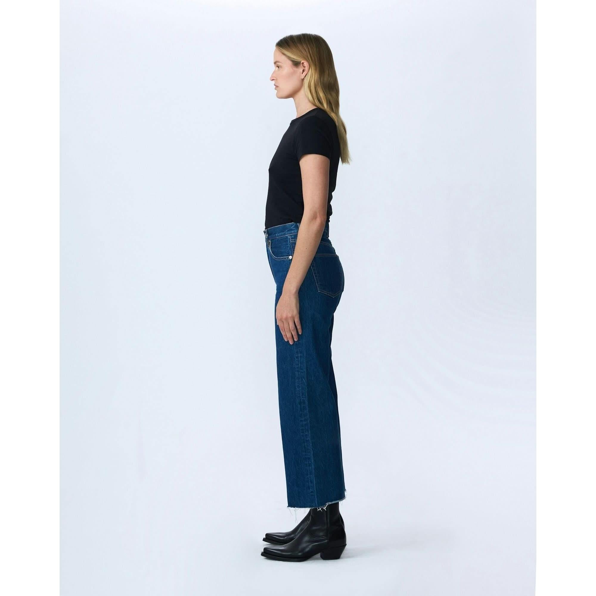 A woman with long blonde hair, wearing a black t-shirt, cropped wide-leg blue high-waisted Slvrlake Denim Grace Crop Revolution jeans from SLVRLAKE, and black ankle boots, is standing in profile view against a plain white background.