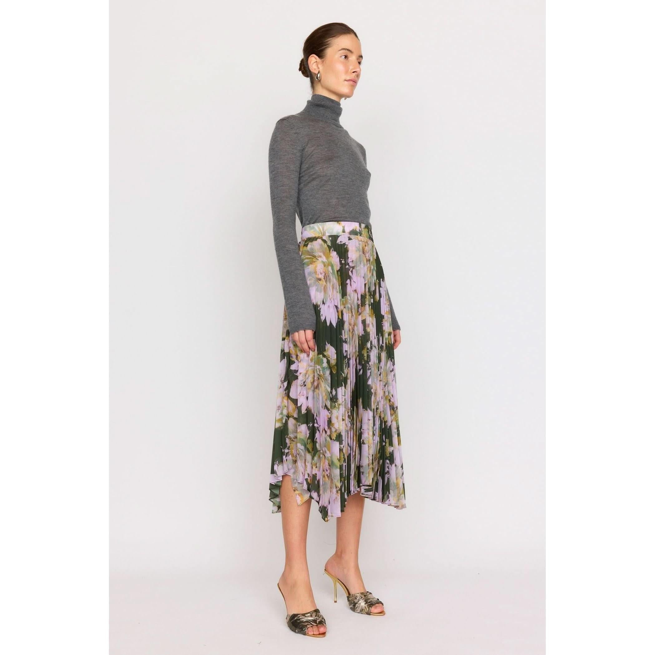 A person wearing a gray turtleneck and the Christy Lynn Gwen Skirt in Midnight Garden, which is a pleated floral skirt in shades of green, pink, and yellow with a fitted waist, stands against a plain white background. They are also wearing patterned high-heeled sandals.