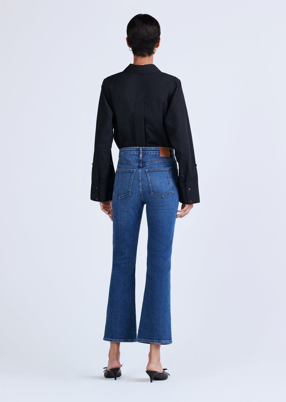 A person with short dark hair is standing with their back to the camera, wearing a black long-sleeved shirt, Derek Lam 10 Crosby High Rise Crop Flare Jeans by Derek Lam in premium denim blue, and black pointed-toe heels. The background is plain white.