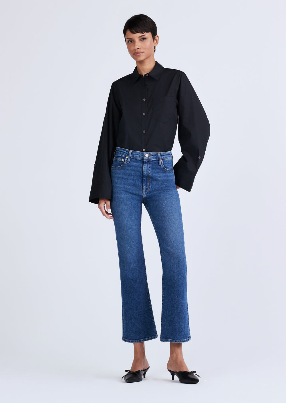 A person with short hair is standing against a plain background, wearing a black long-sleeve button-up shirt and Derek Lam 10 Crosby High Rise Crop Flare Jeans. They have on black pointed-toe shoes with a slight heel. The outfit is casual yet sophisticated.