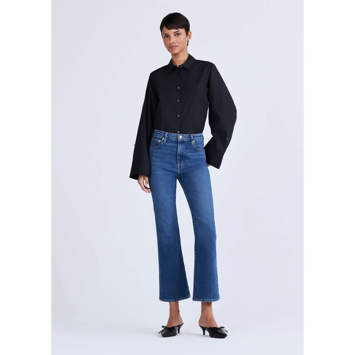 A person with short hair is standing against a plain background, wearing a black long-sleeve button-up shirt and Derek Lam 10 Crosby High Rise Crop Flare Jeans. They have on black pointed-toe shoes with a slight heel. The outfit is casual yet sophisticated.